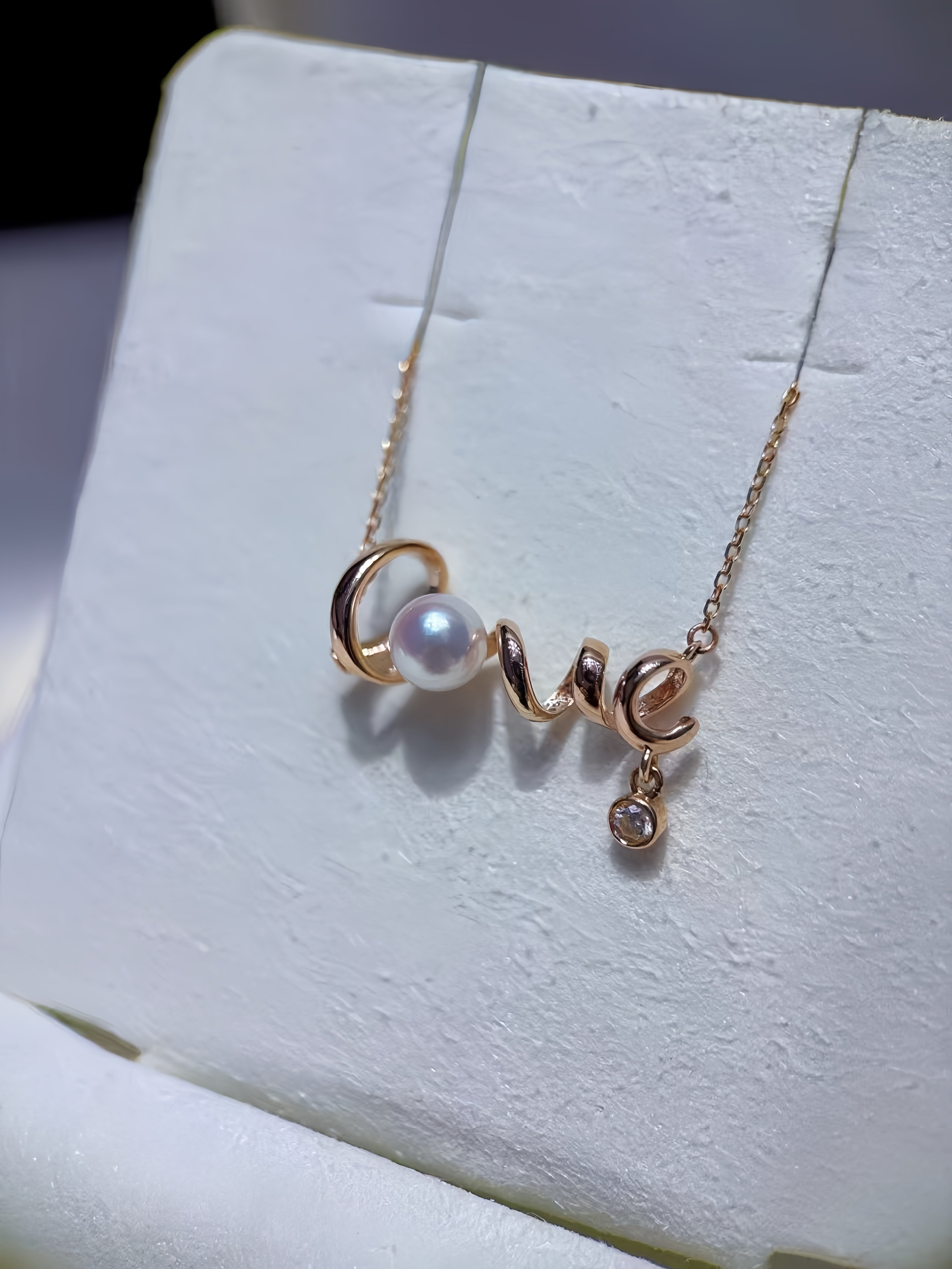 A delicate gold necklace with a script-style pendant spelling 'LOVE', featuring a white pearl and a small clear stone dangle, displayed on a white jewelry stand.