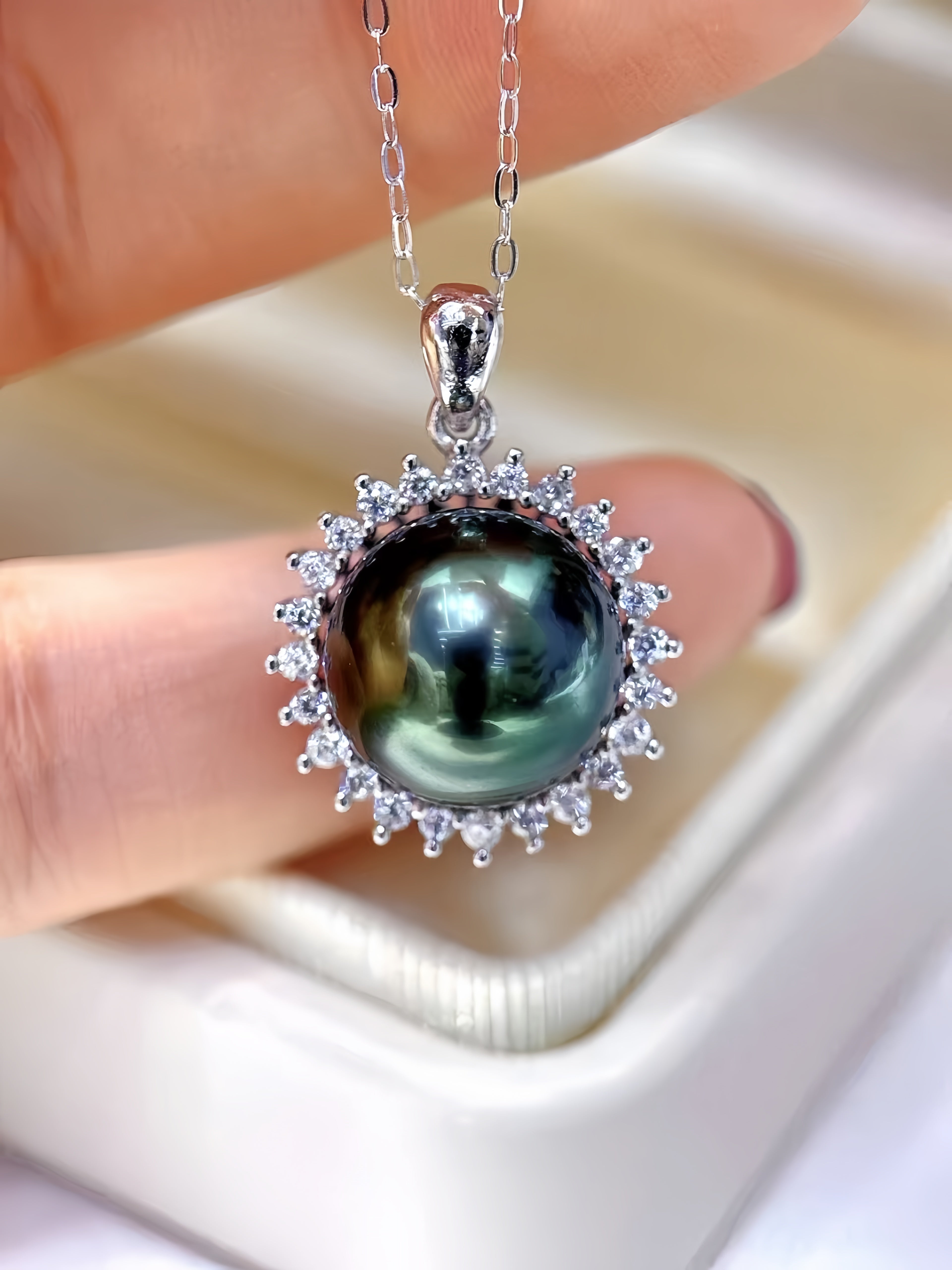 A close-up of a silver pendant necklace held between fingers. The pendant features a large, round, dark green pearl-like stone surrounded by a halo of small clear stones, suspended from a silver chain.