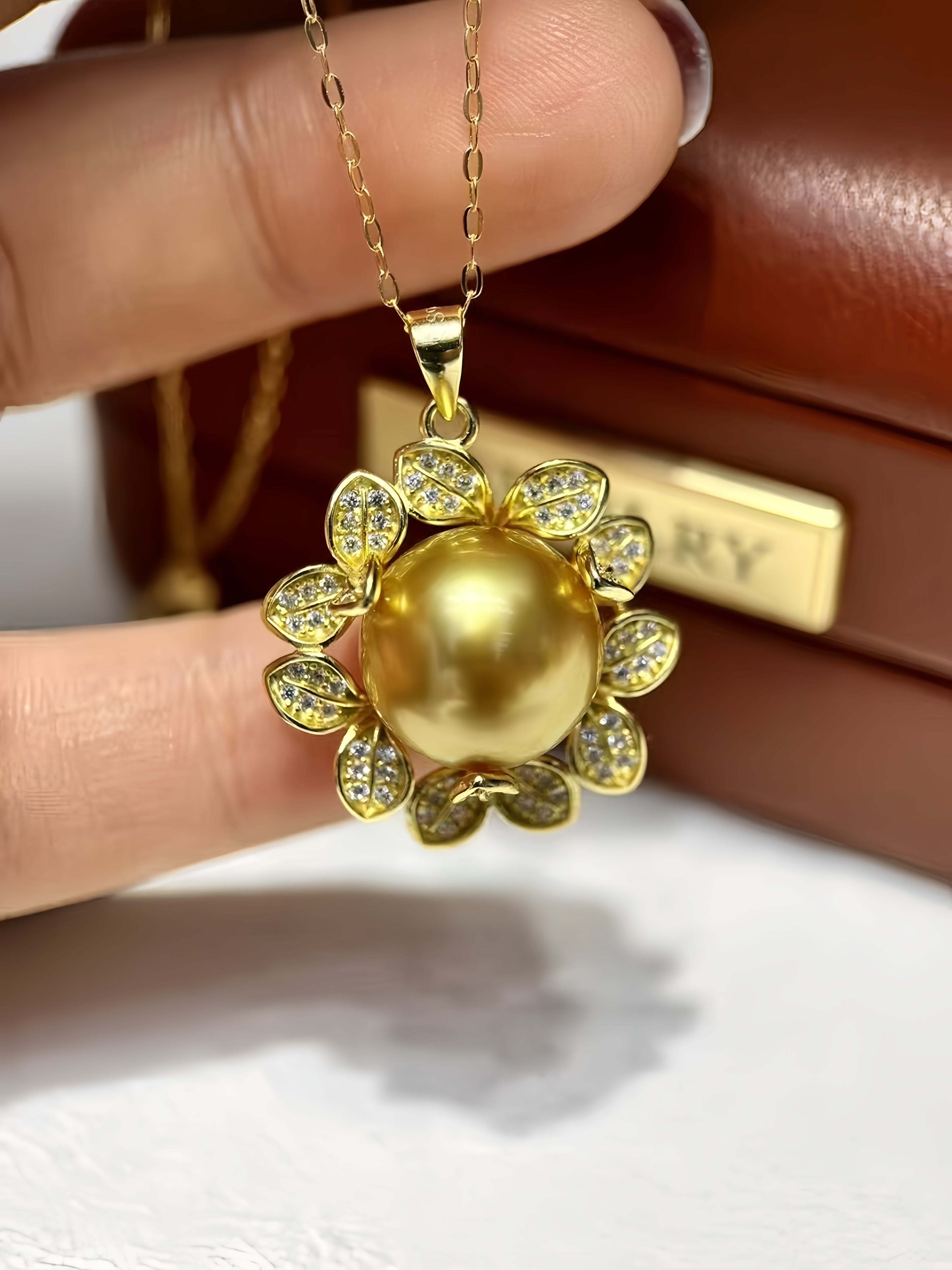 A gold pendant necklace featuring a large round pearl-like center surrounded by small clear stones in a floral petal design, held by fingers in front of a brown leather box.