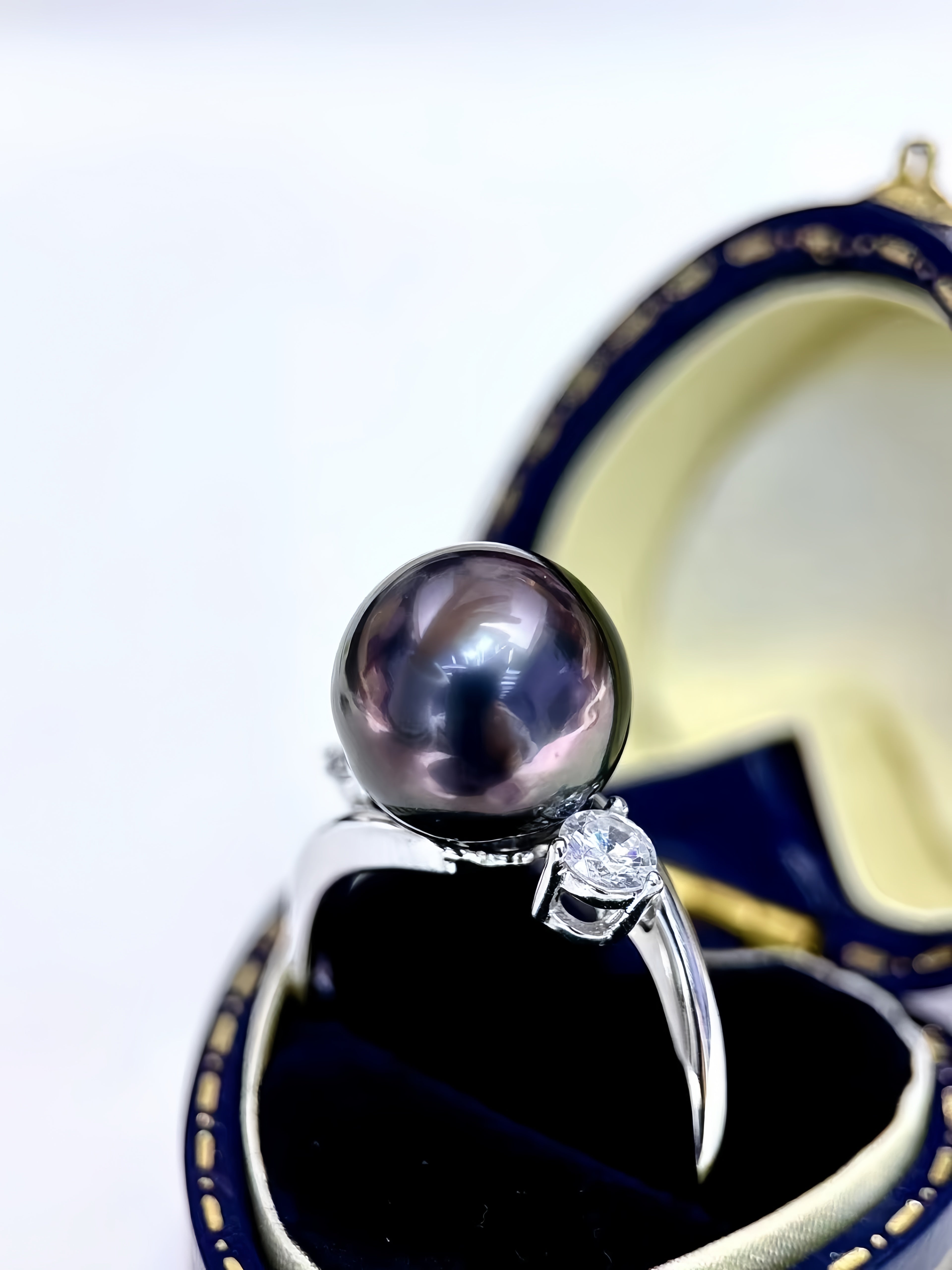 A silver ring featuring a large, lustrous dark purple pearl and a small clear gemstone, displayed on a blue and cream ring box.
