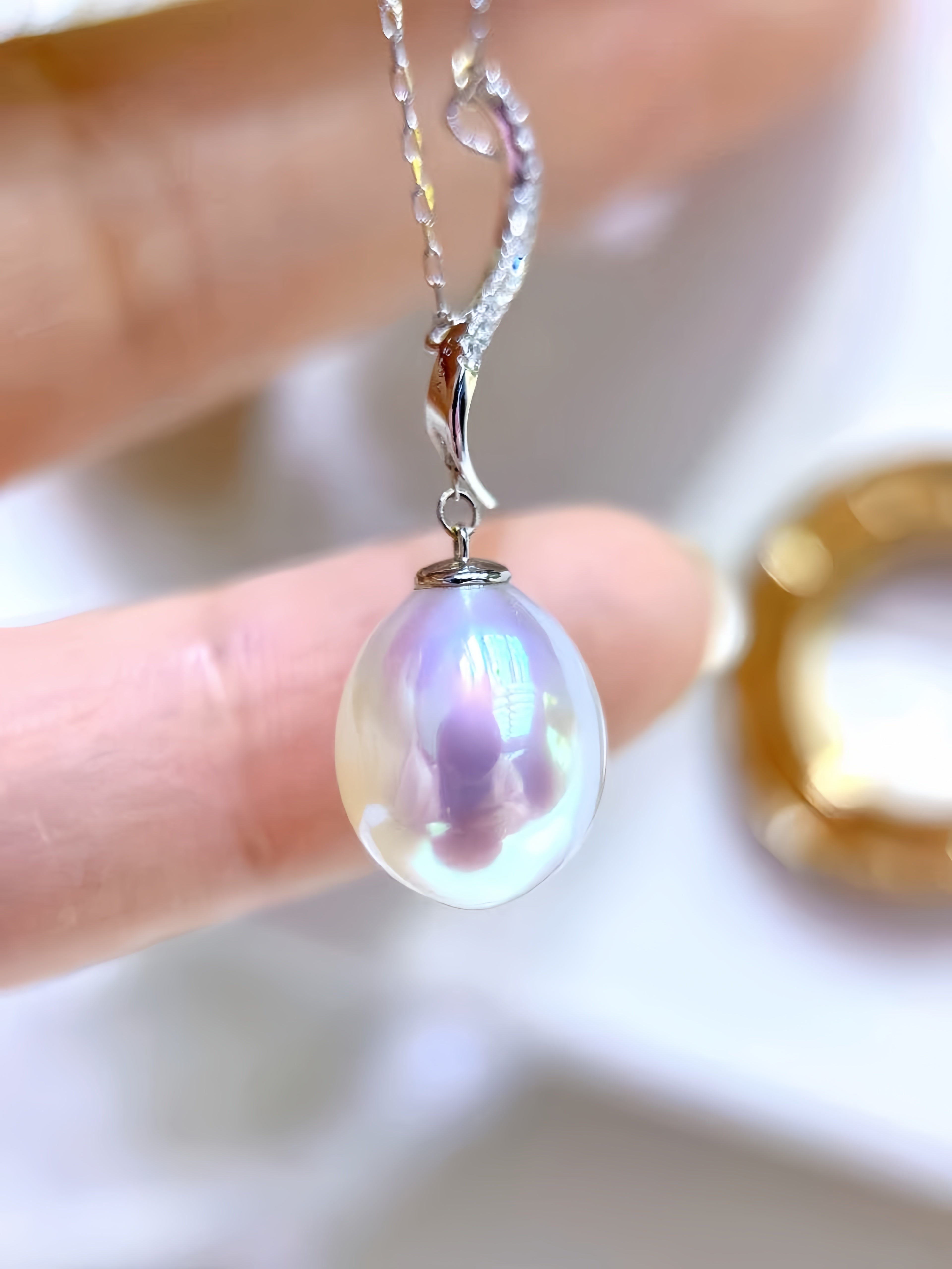 A close-up of a silver necklace with a teardrop-shaped iridescent pearl pendant, held by fingers against a soft, blurred background.