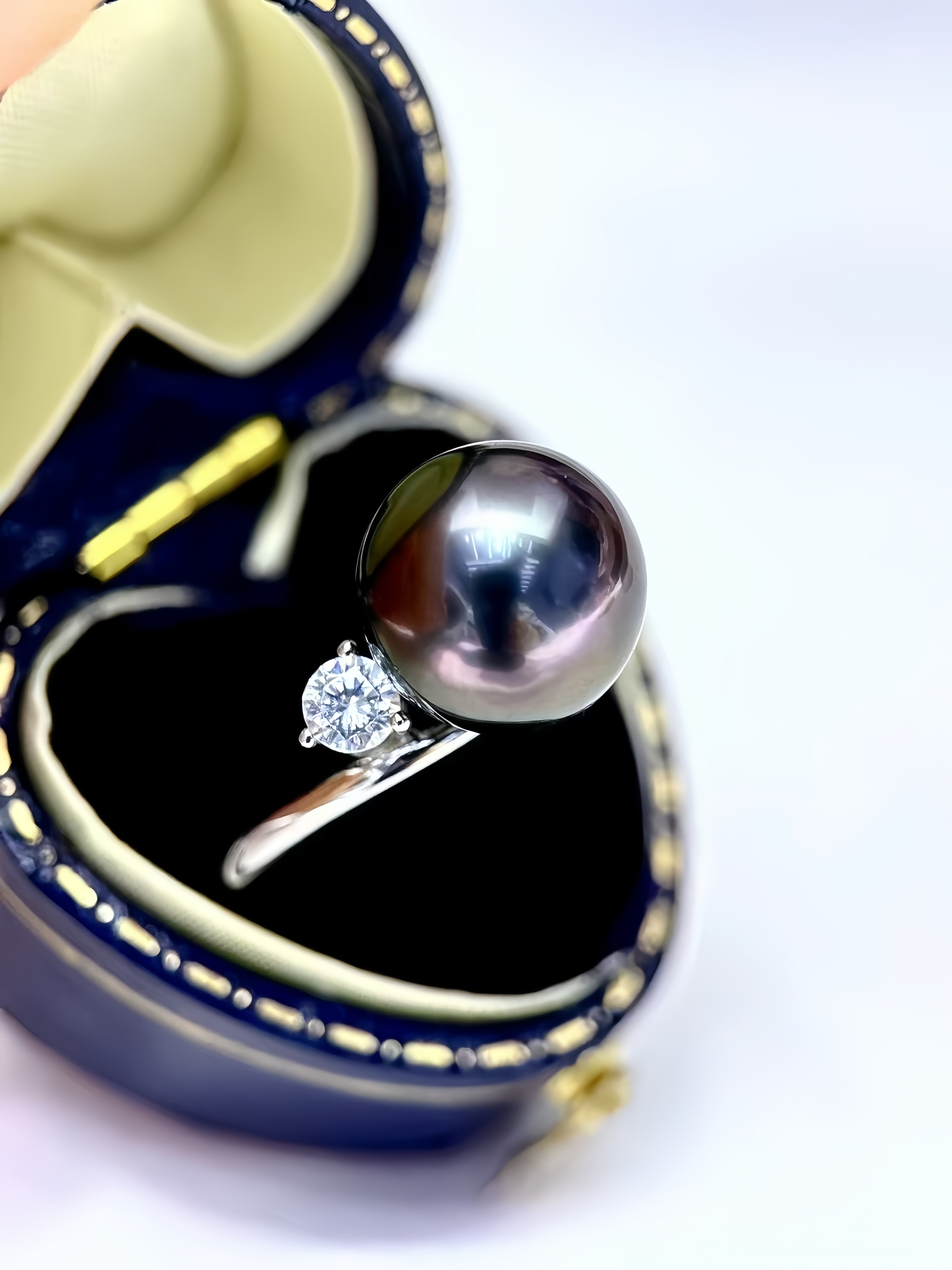 A close-up of a pearl ring displayed in an open navy blue jewelry box. The ring features a large, dark gray-black round pearl on a silver band with a small clear diamond accent.