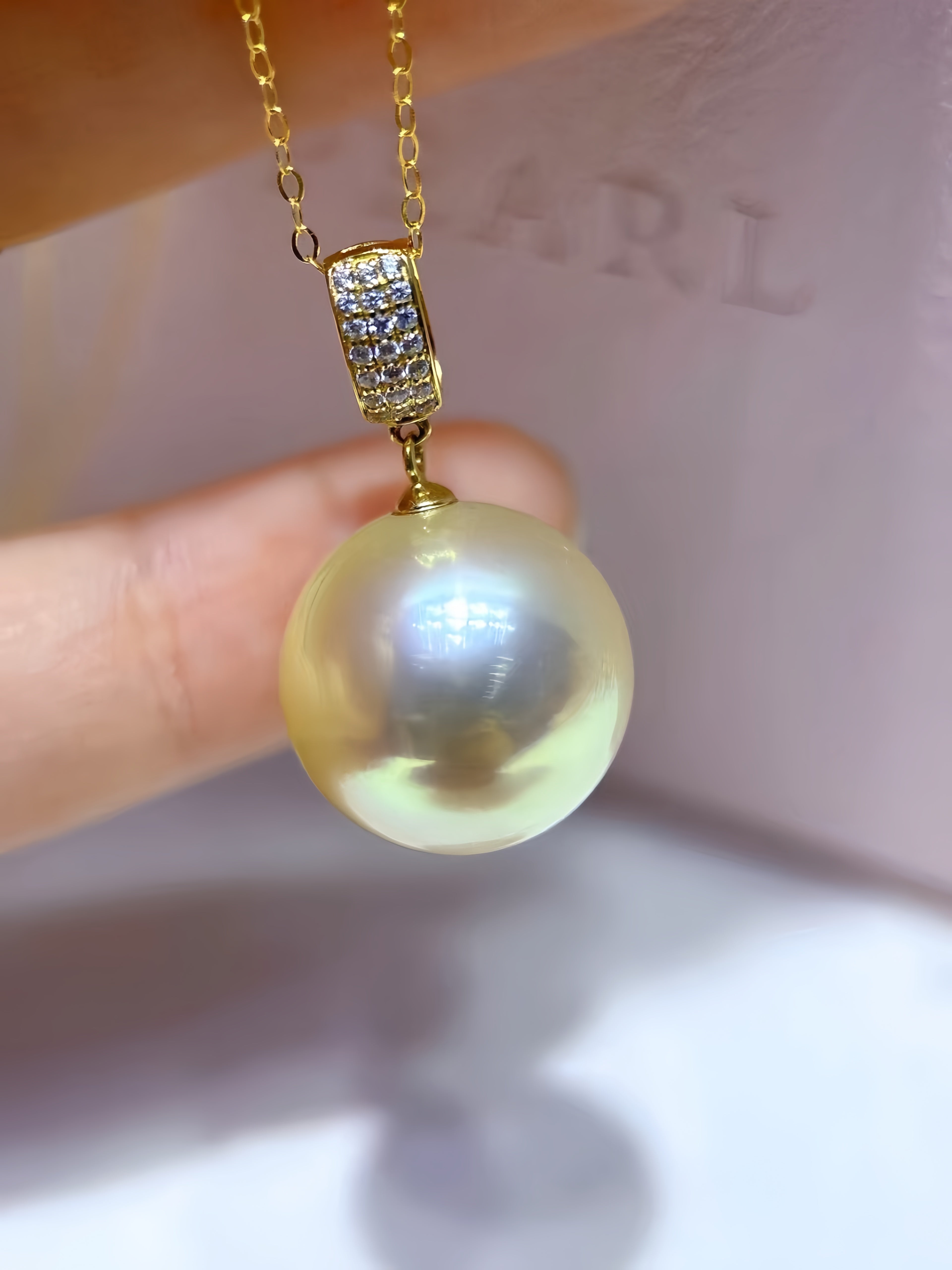 A close-up of a gold pendant necklace featuring a large, lustrous pearl pendant and a gold bail set with small clear stones, held by fingers against a soft pink background.