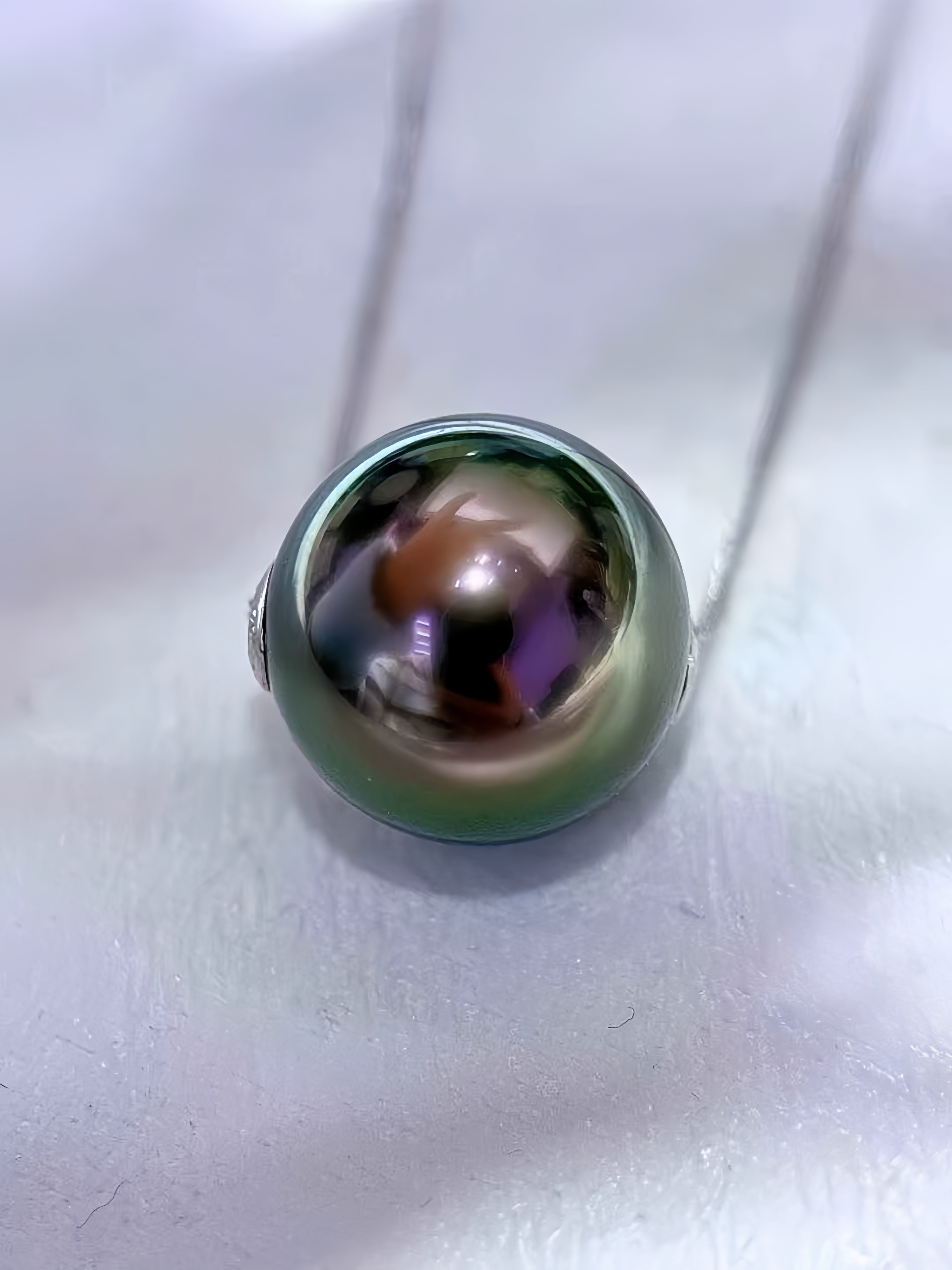 A close-up of a round freshwater pearl pendant on a silver chain, resting on a textured white surface.