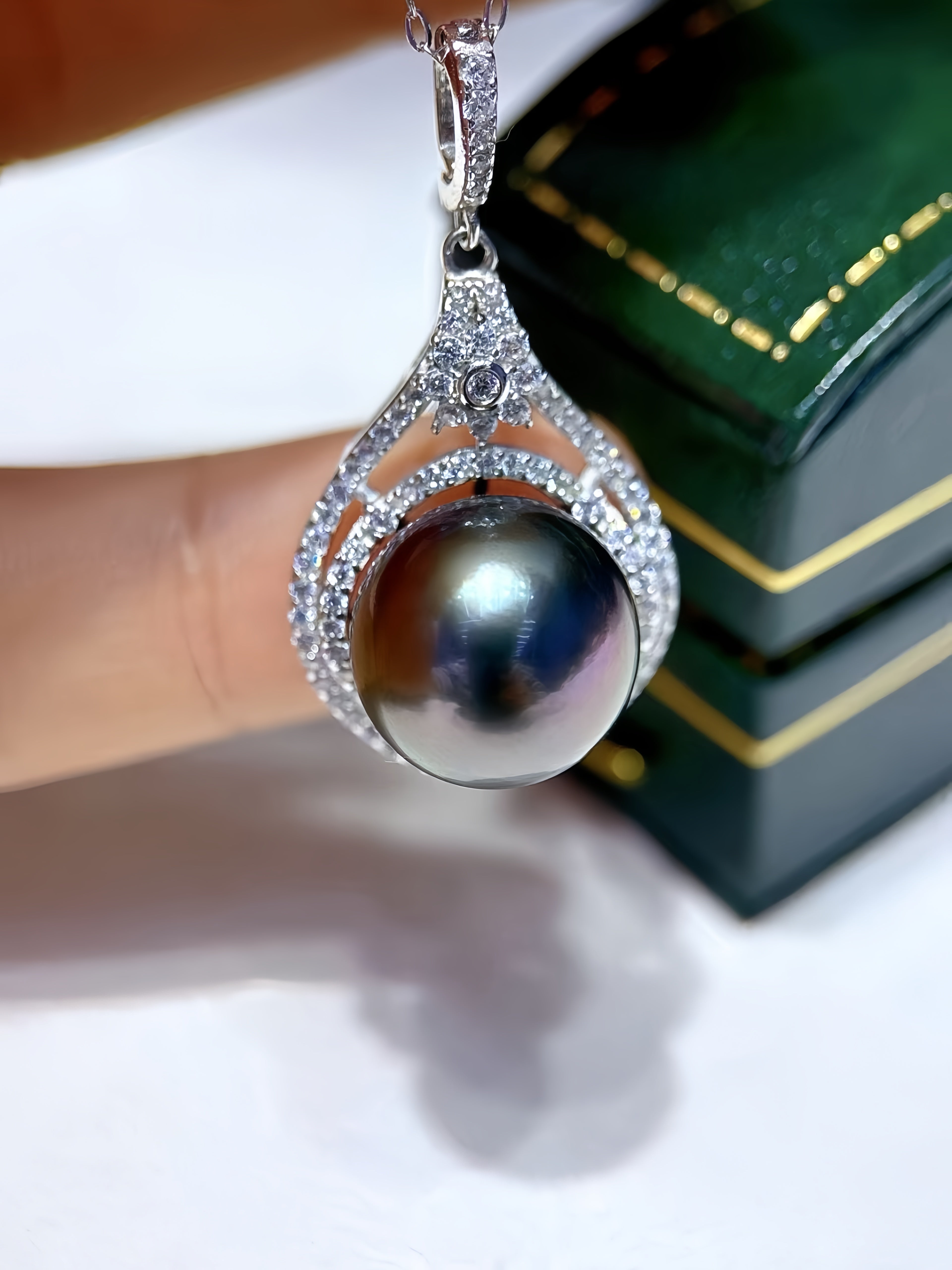 A close-up of a teardrop-shaped pendant featuring a large iridescent dark pearl surrounded by silver-toned pavé-set stones, held near a green jewelry box.