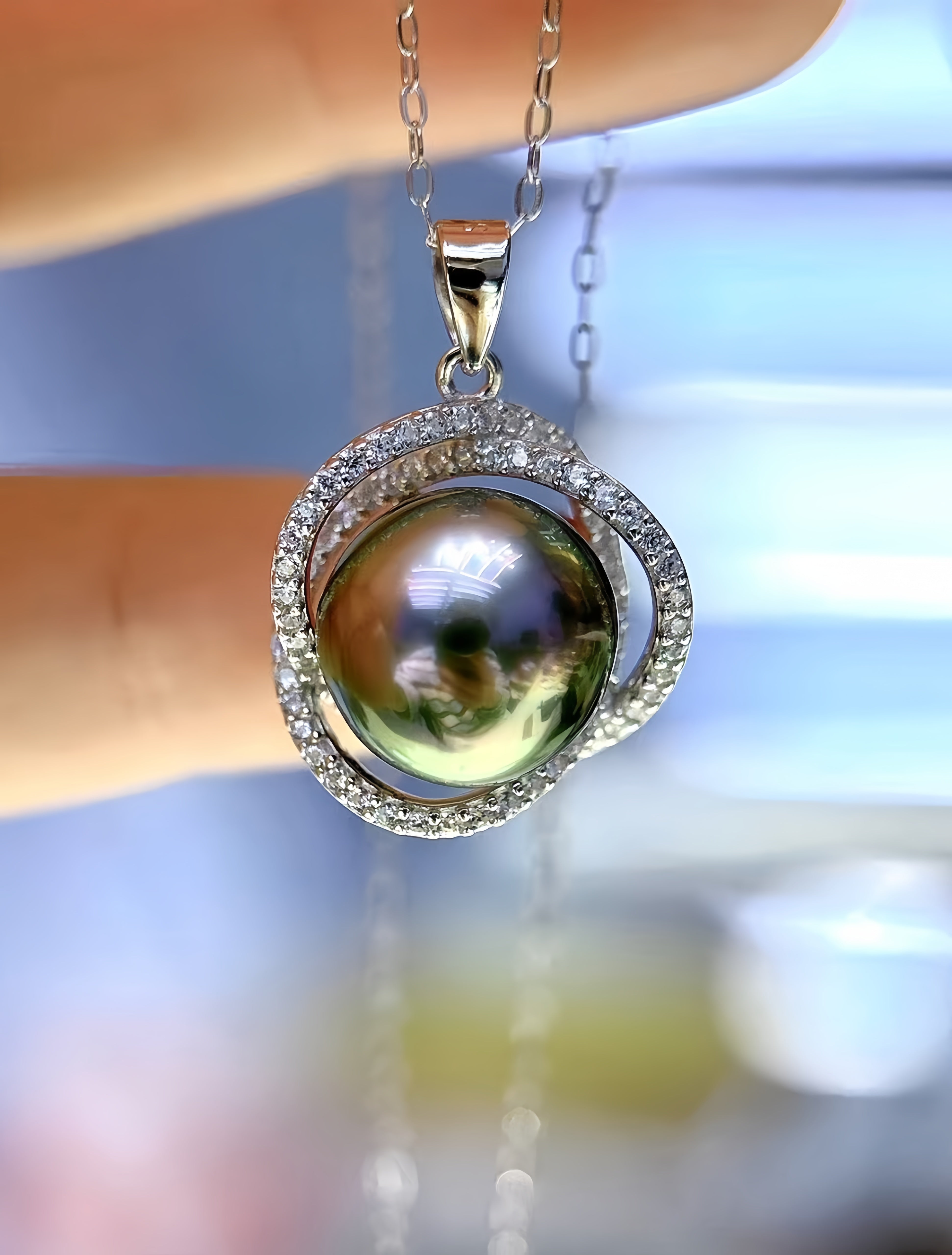 A close-up of a silver pendant necklace with a large iridescent green pearl surrounded by a halo of small clear stones, held between fingers against a blurred light blue background.