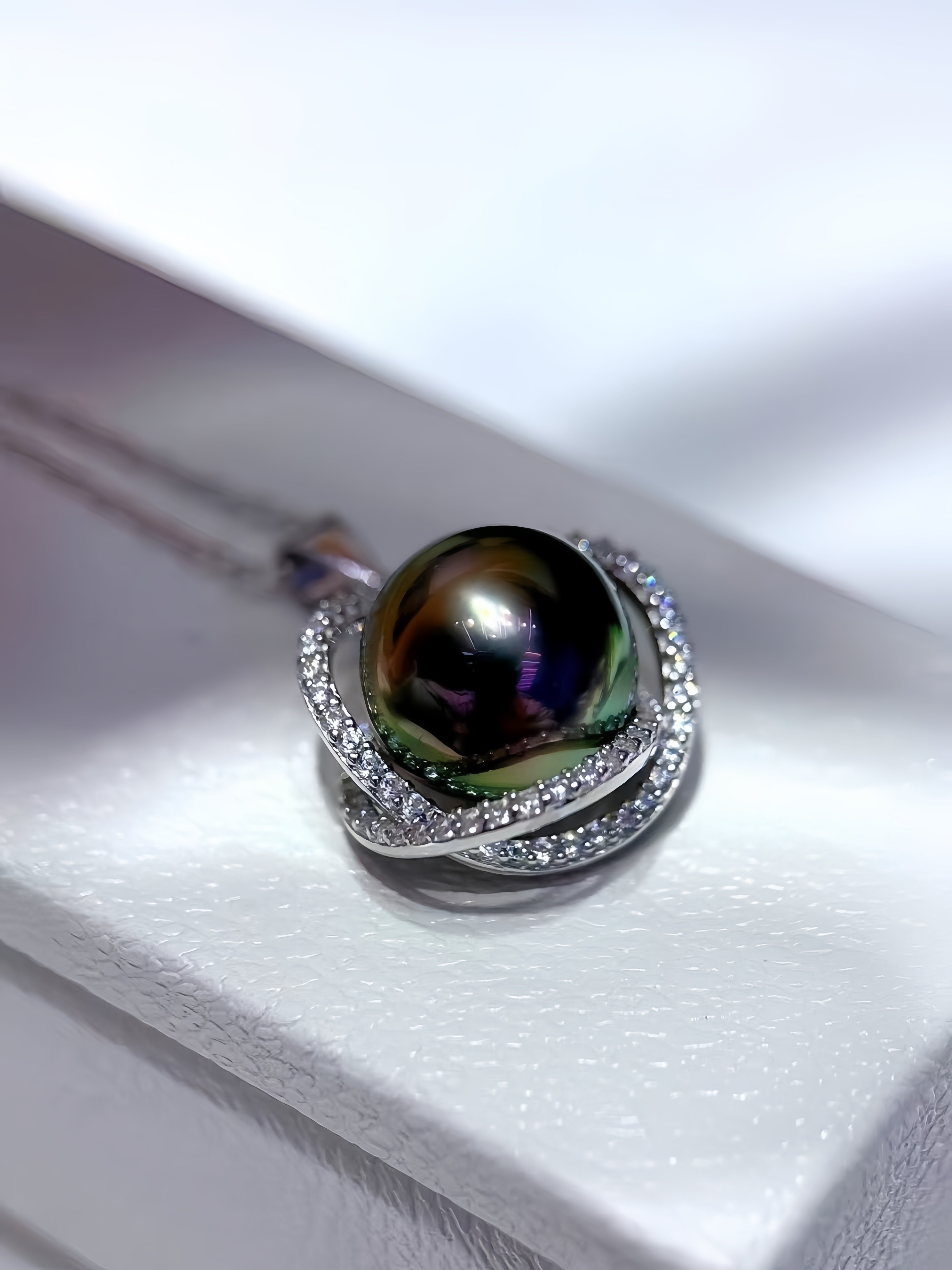 A close-up of a pendant necklace featuring a large, round, iridescent black pearl surrounded by silver-toned pavé-set accents, resting on a white textured jewelry box.