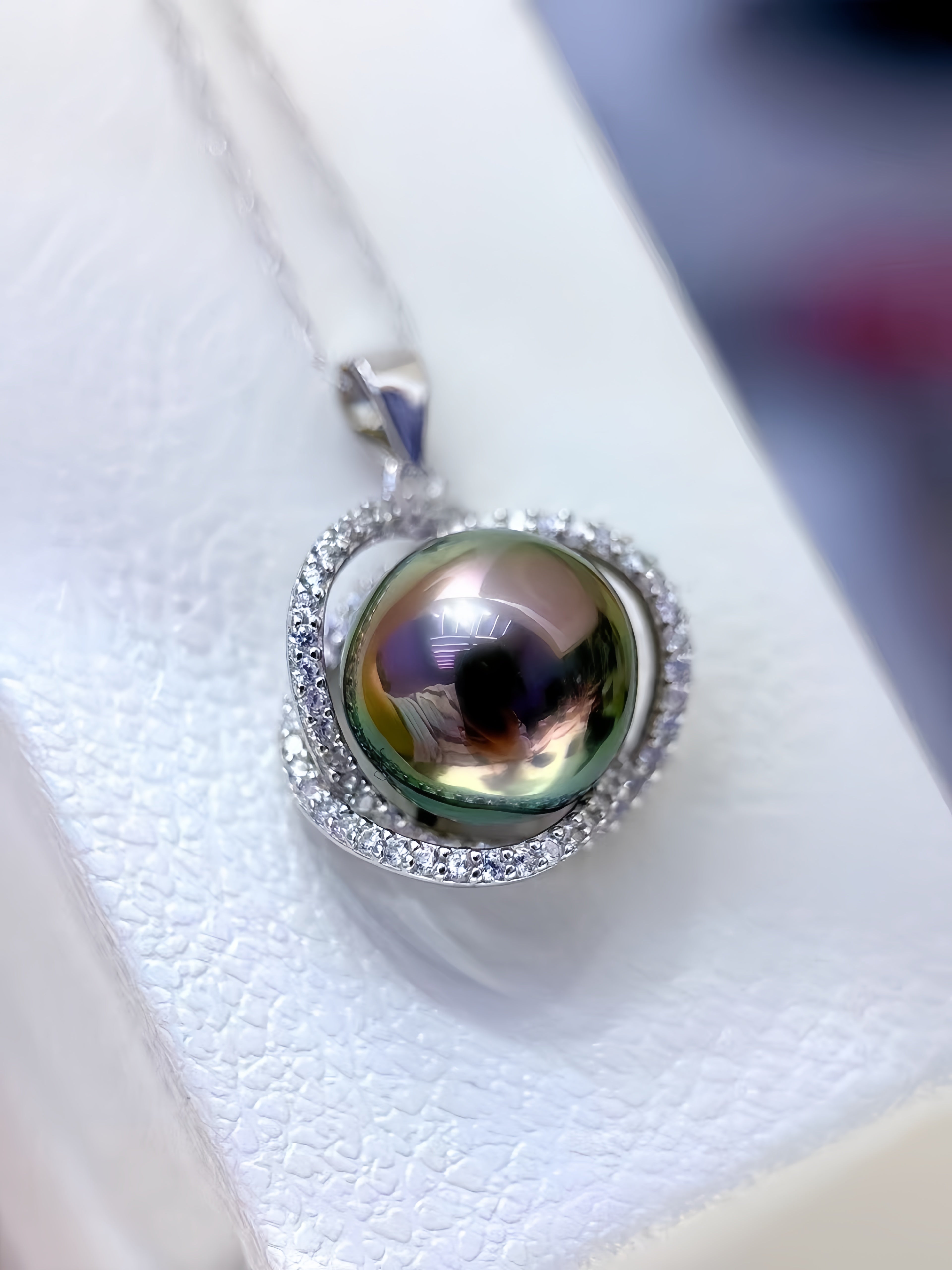 A close-up of a silver pendant necklace featuring a large, iridescent green pearl surrounded by a halo of small clear stones, resting on a textured white surface.