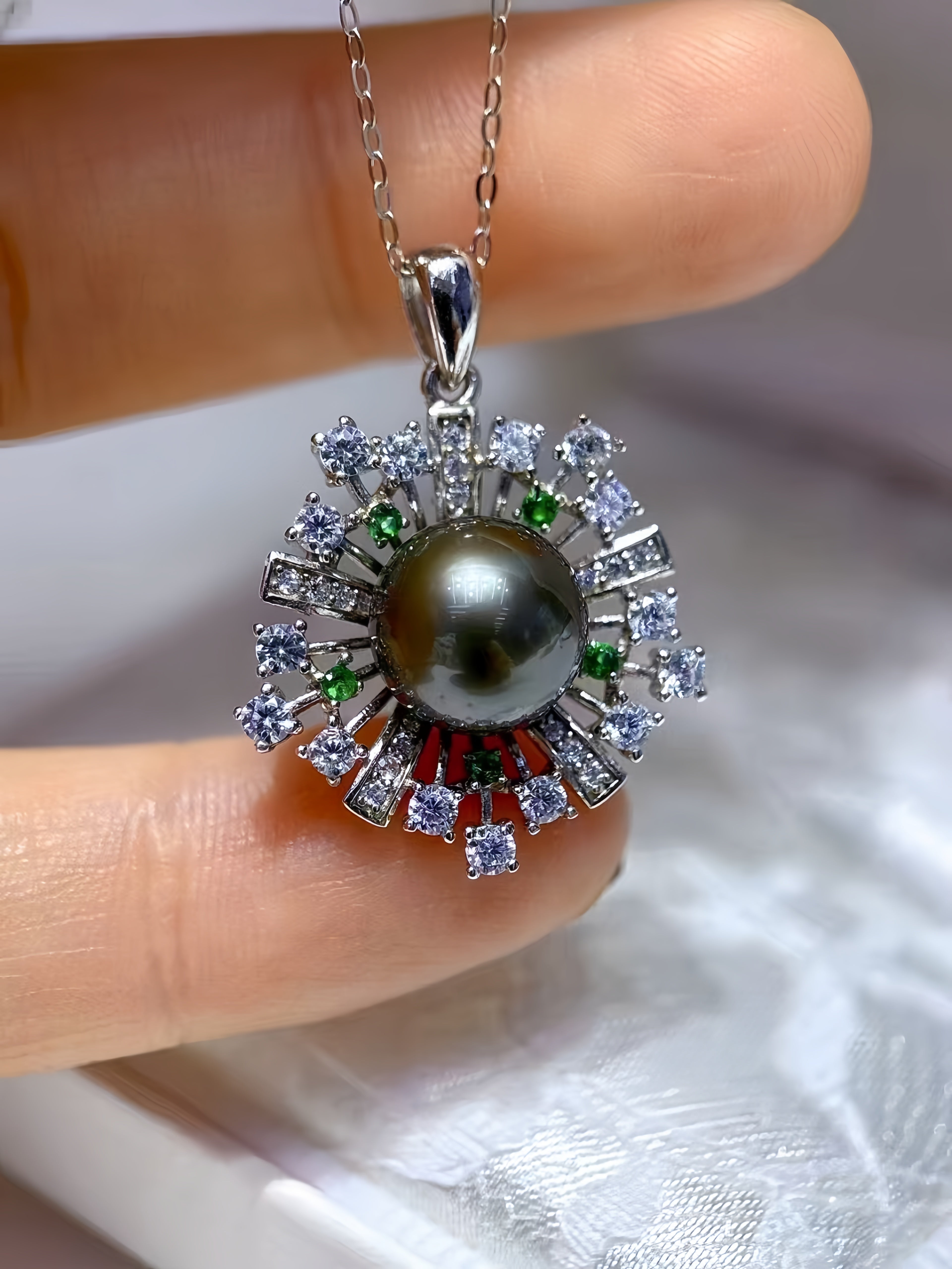 A close-up of a pendant necklace held between fingers. The pendant features a large dark gray pearl surrounded by silver-tone rays set with clear stones, small green and red accent stones, and a delicate silver chain.