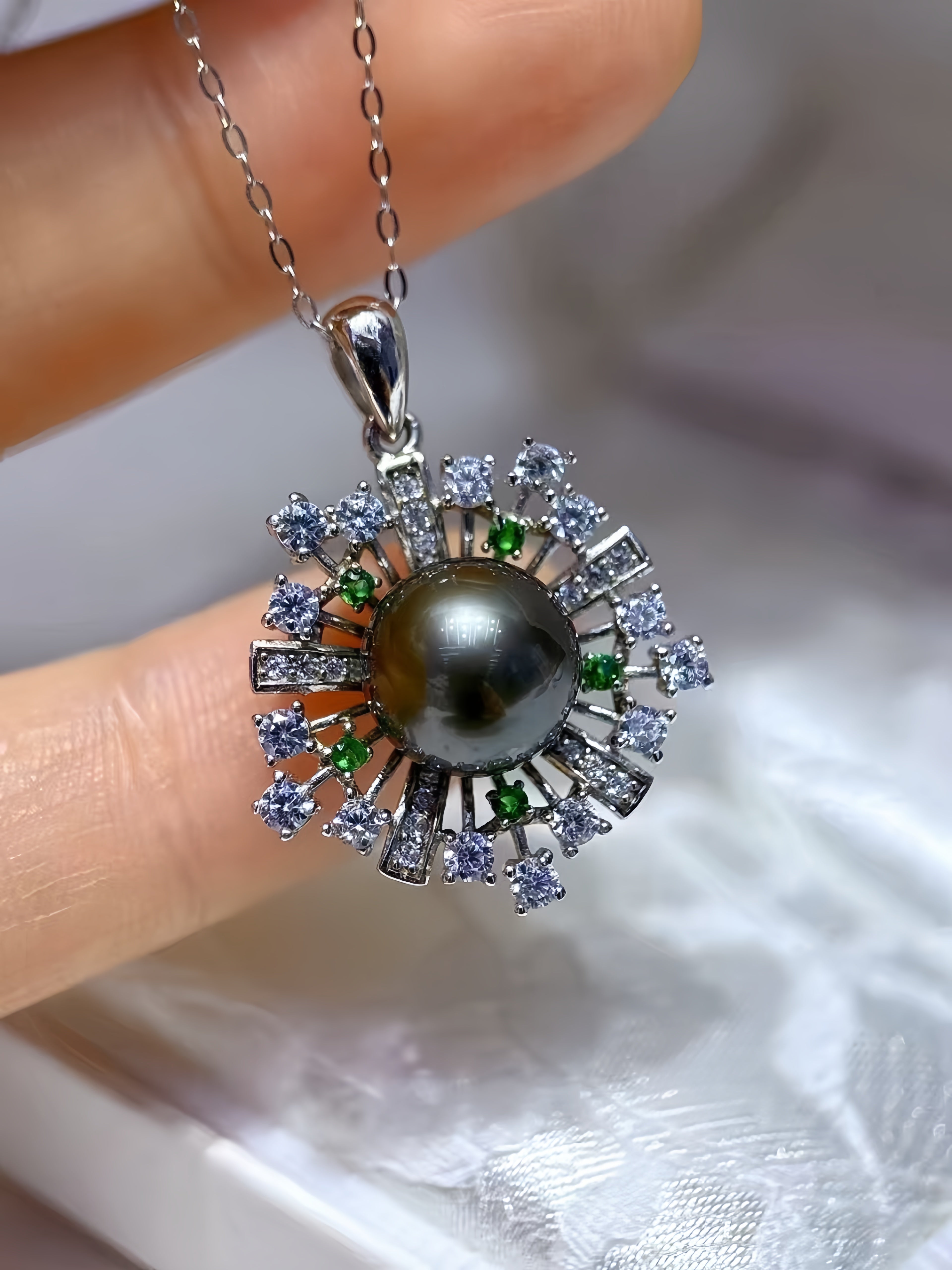 A close-up of a hand holding a silver pendant necklace featuring a dark gray pearl surrounded by clear stones and small green accents, with a white satin-like background.