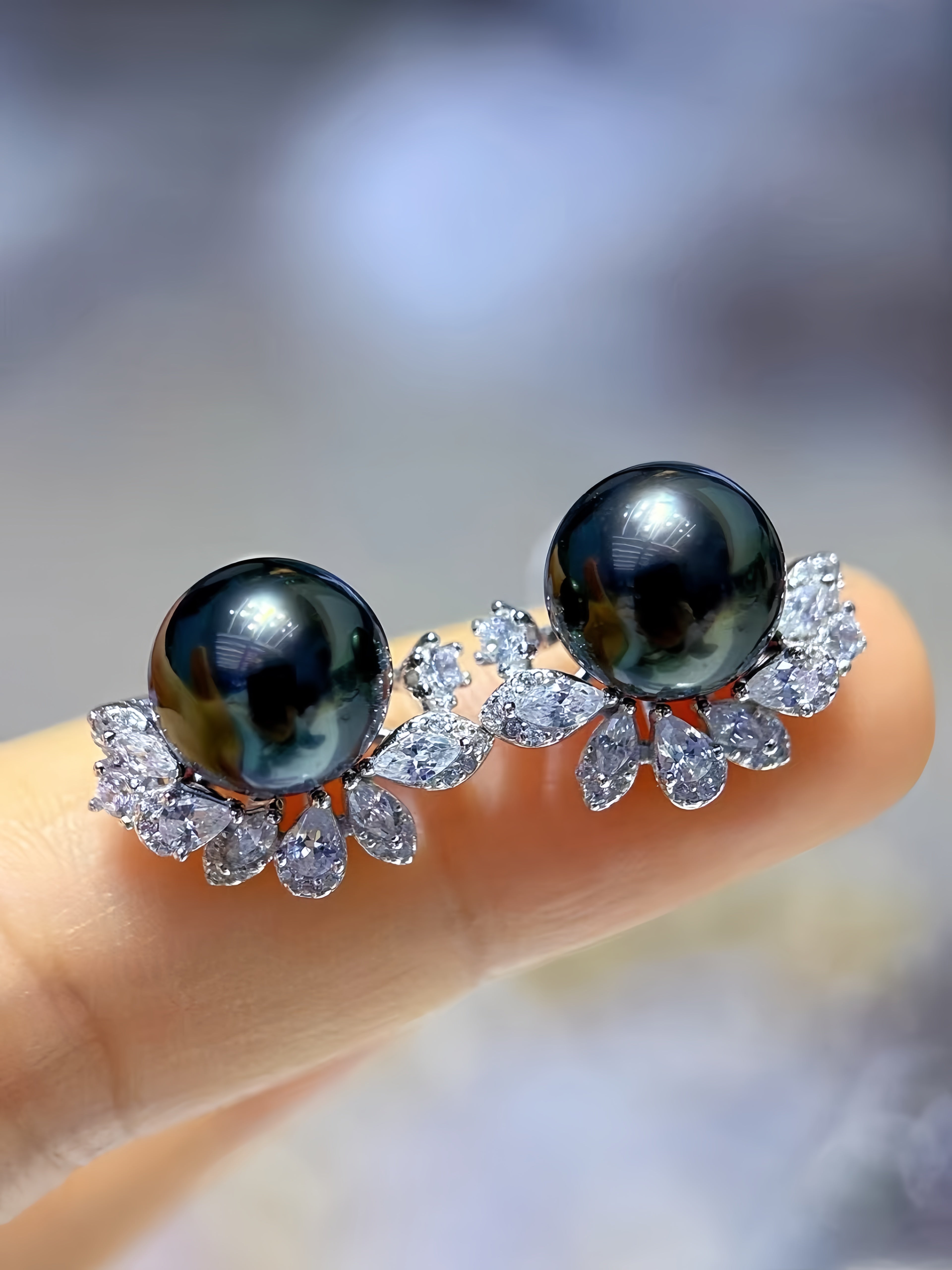 A pair of stud earrings with large round black pearls and clear teardrop-shaped stones, shown resting on a person's finger against a soft blurred background.