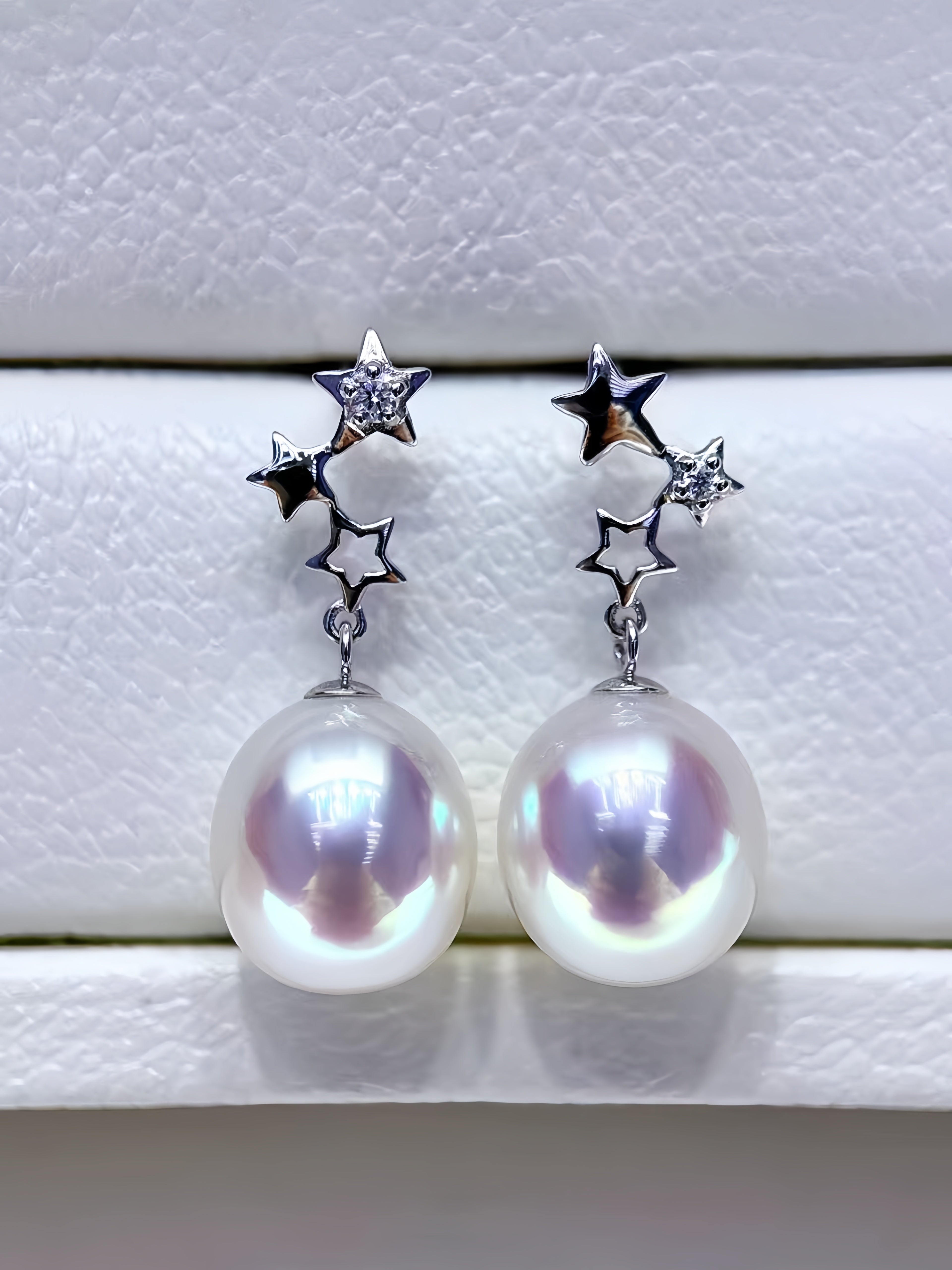 A pair of silver star cluster drop earrings with large iridescent white freshwater pearls, displayed on a textured white jewelry box.