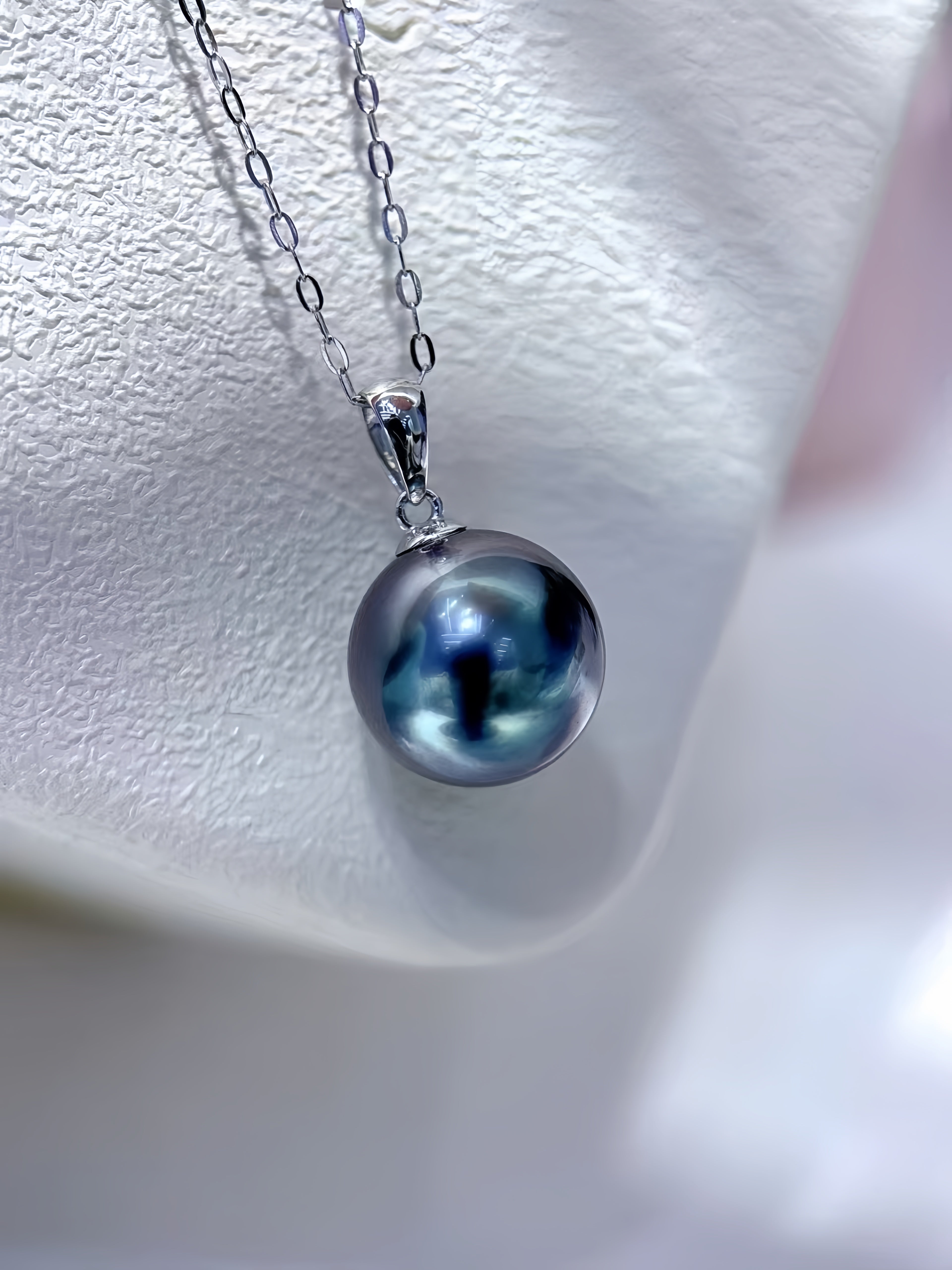 A silver pendant necklace featuring a large, round, iridescent dark blue-gray pearl on a delicate silver chain, displayed on a textured white jewelry stand.