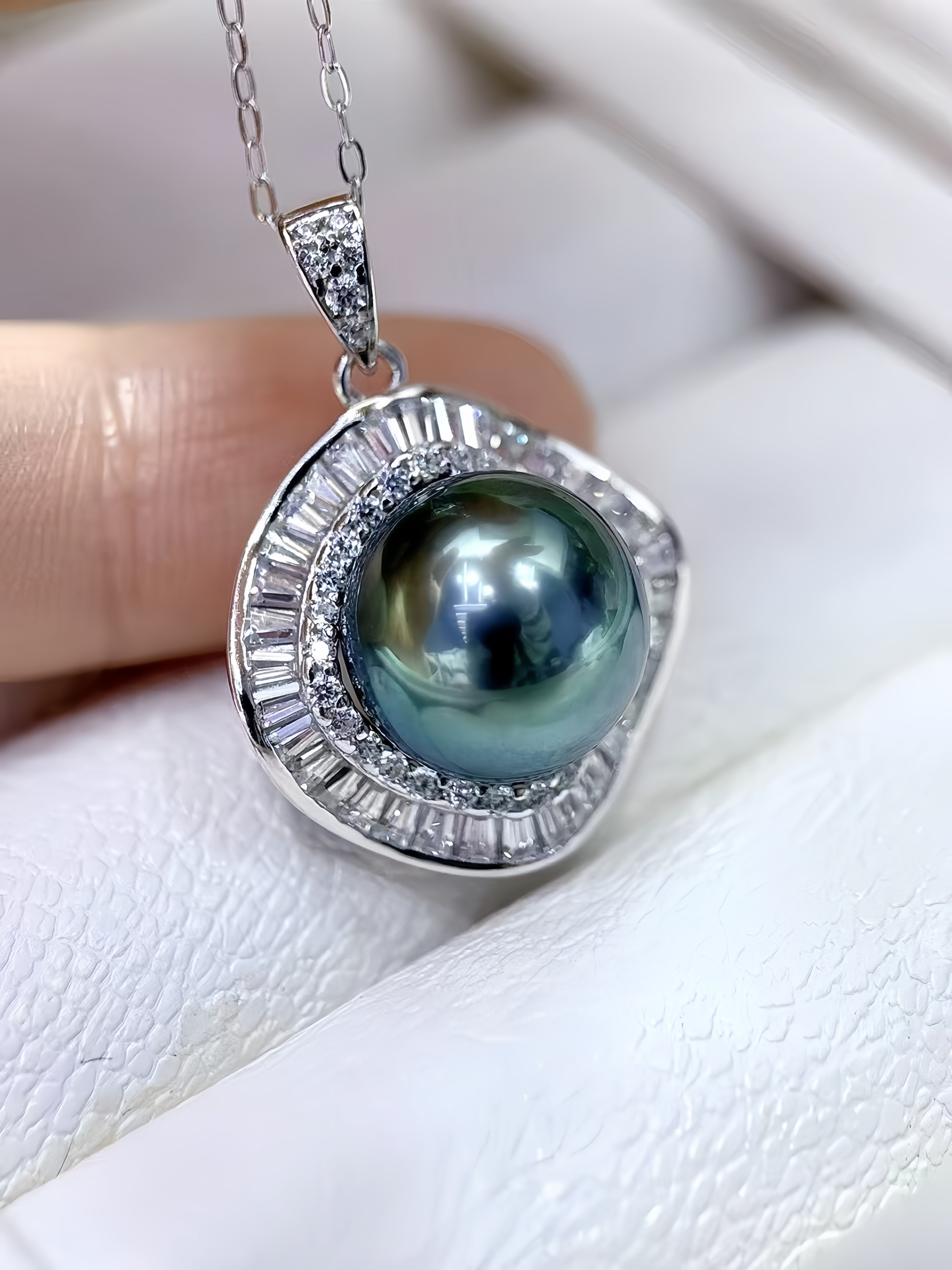 A close-up of a silver pendant necklace featuring a round iridescent dark blue-green pearl surrounded by a halo of clear baguette and round stones, held against a white textured background.