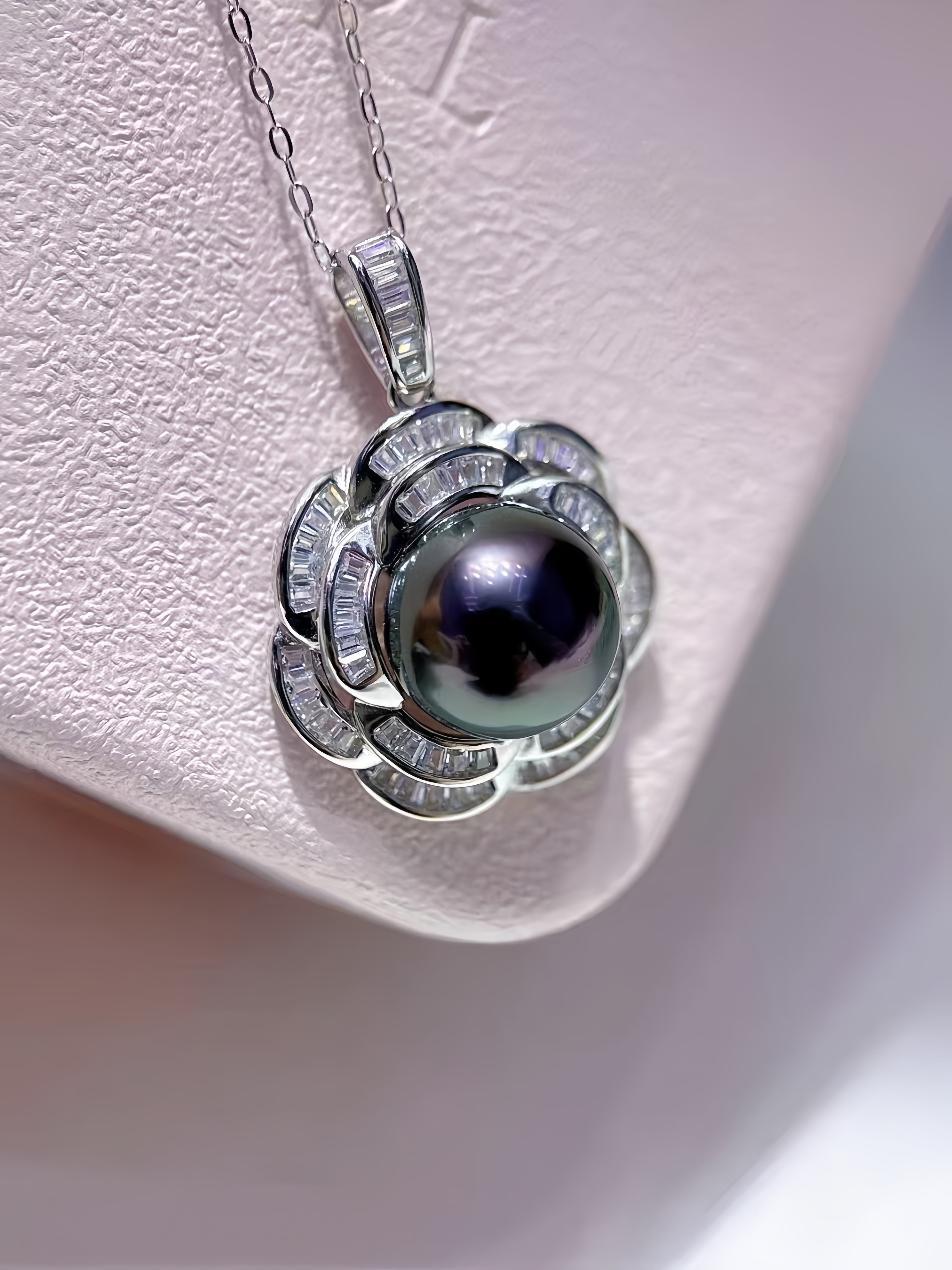 A silver pendant necklace featuring a central iridescent dark pearl surrounded by a flower-like halo of small clear stones, displayed on a textured light pink jewelry stand.
