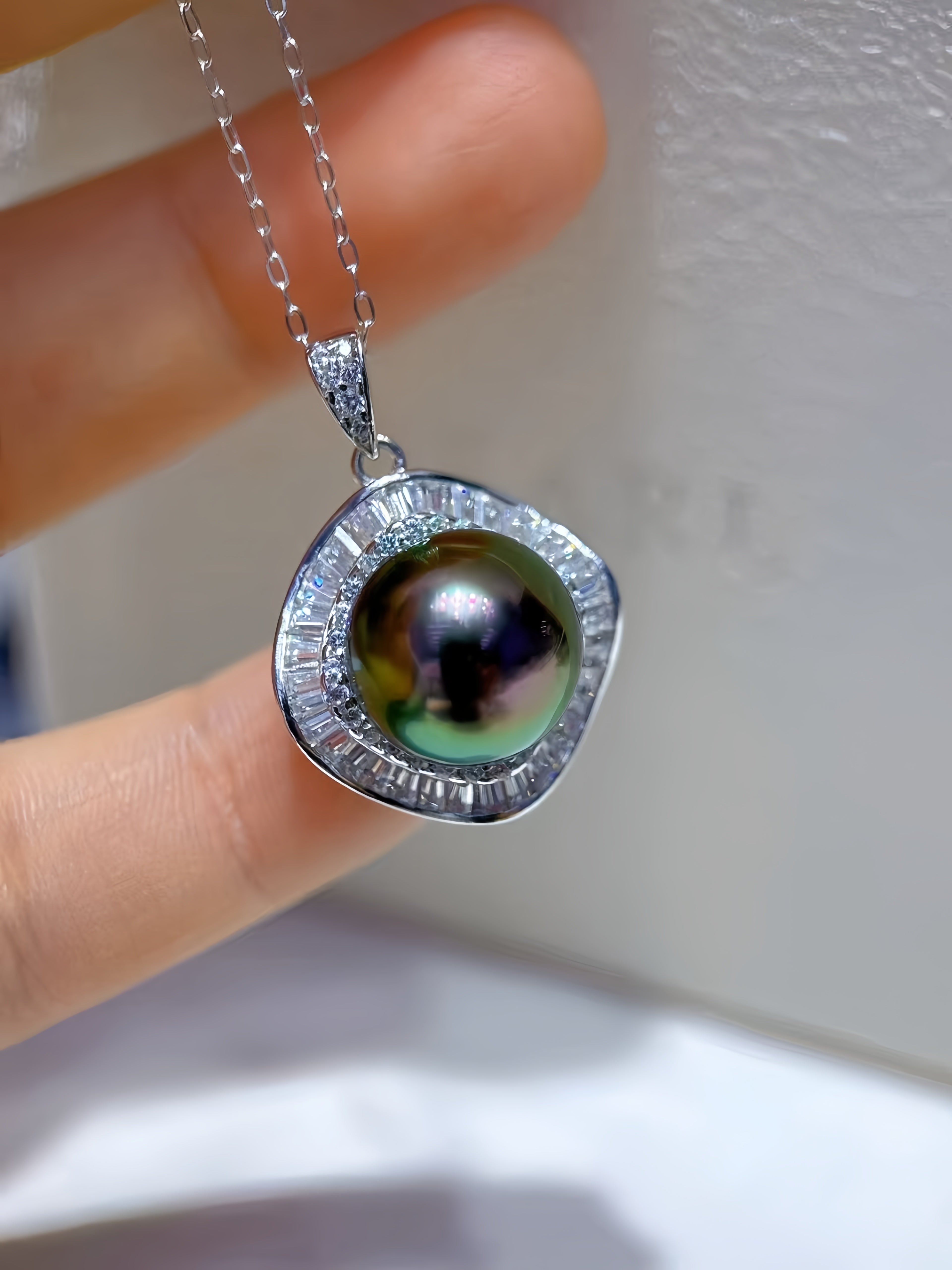 A silver pendant necklace featuring a large, iridescent green pearl surrounded by small clear stones, held by a hand against a light background.