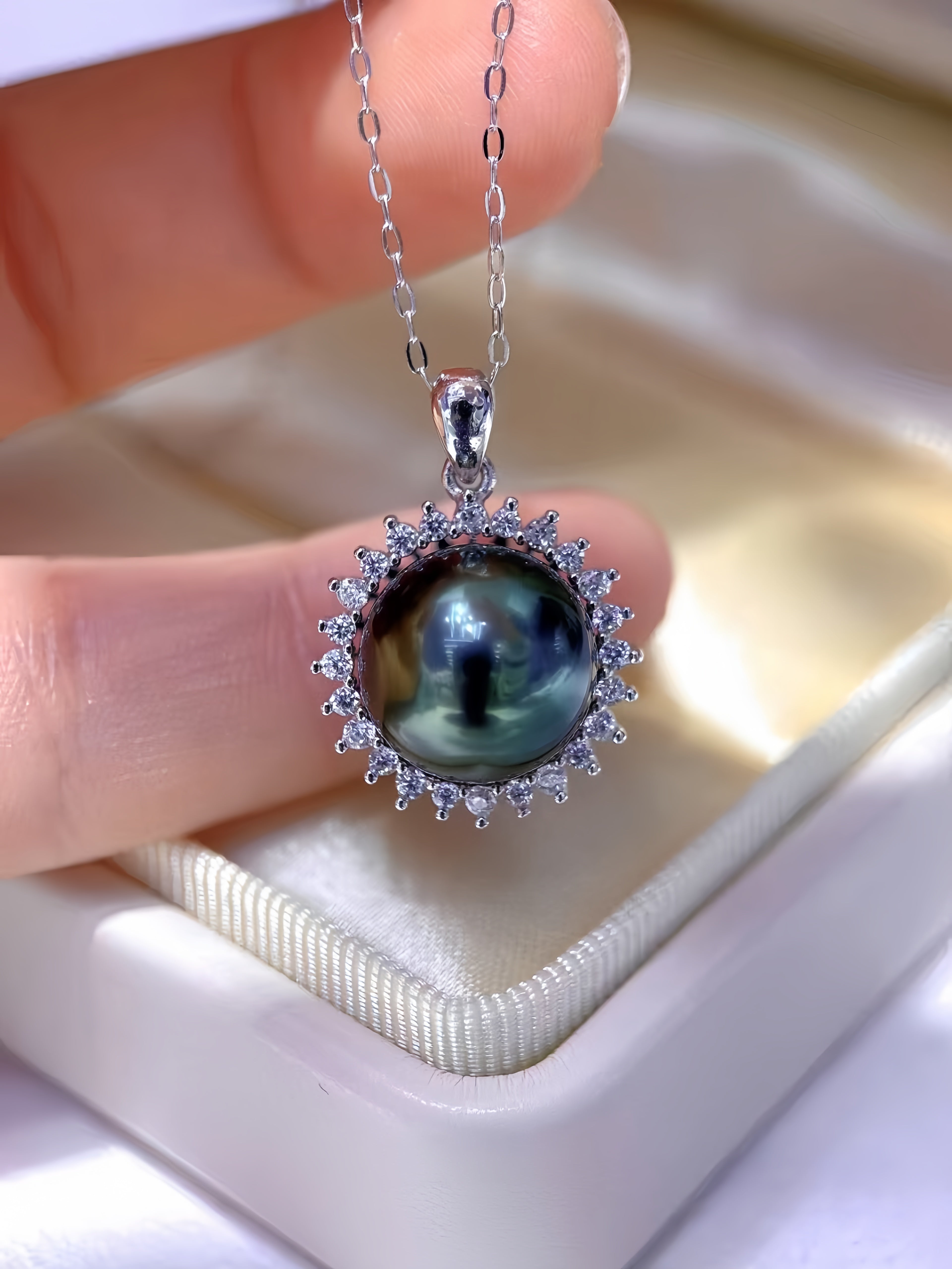 A close-up of a hand holding a silver pendant necklace with a round iridescent dark pearl surrounded by small clear stones, resting over an open cream-colored jewelry box.