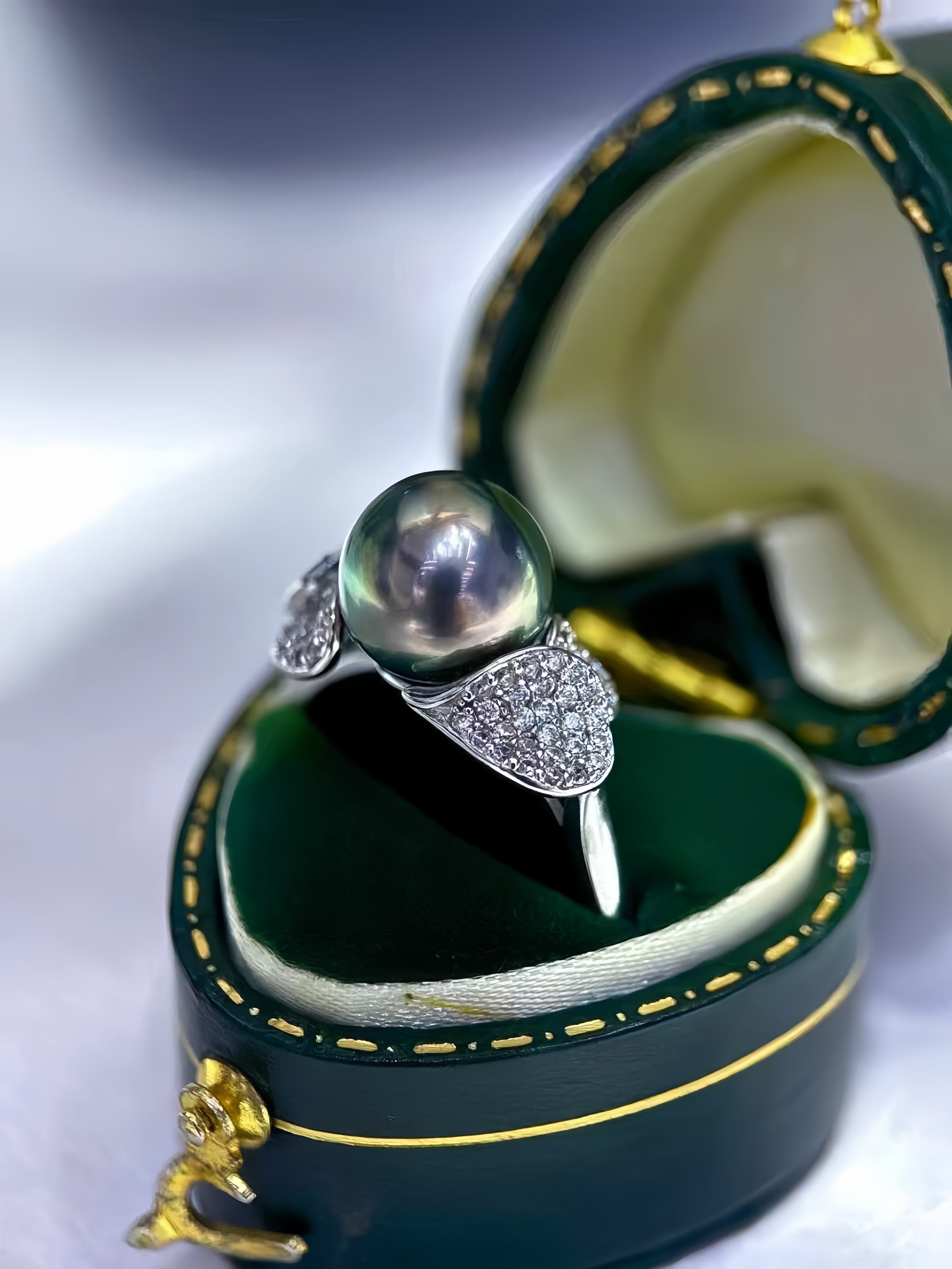 A silver ring with a large dark pearl and pavé-set heart-shaped accents, displayed in an open dark green jewelry box with gold trim.