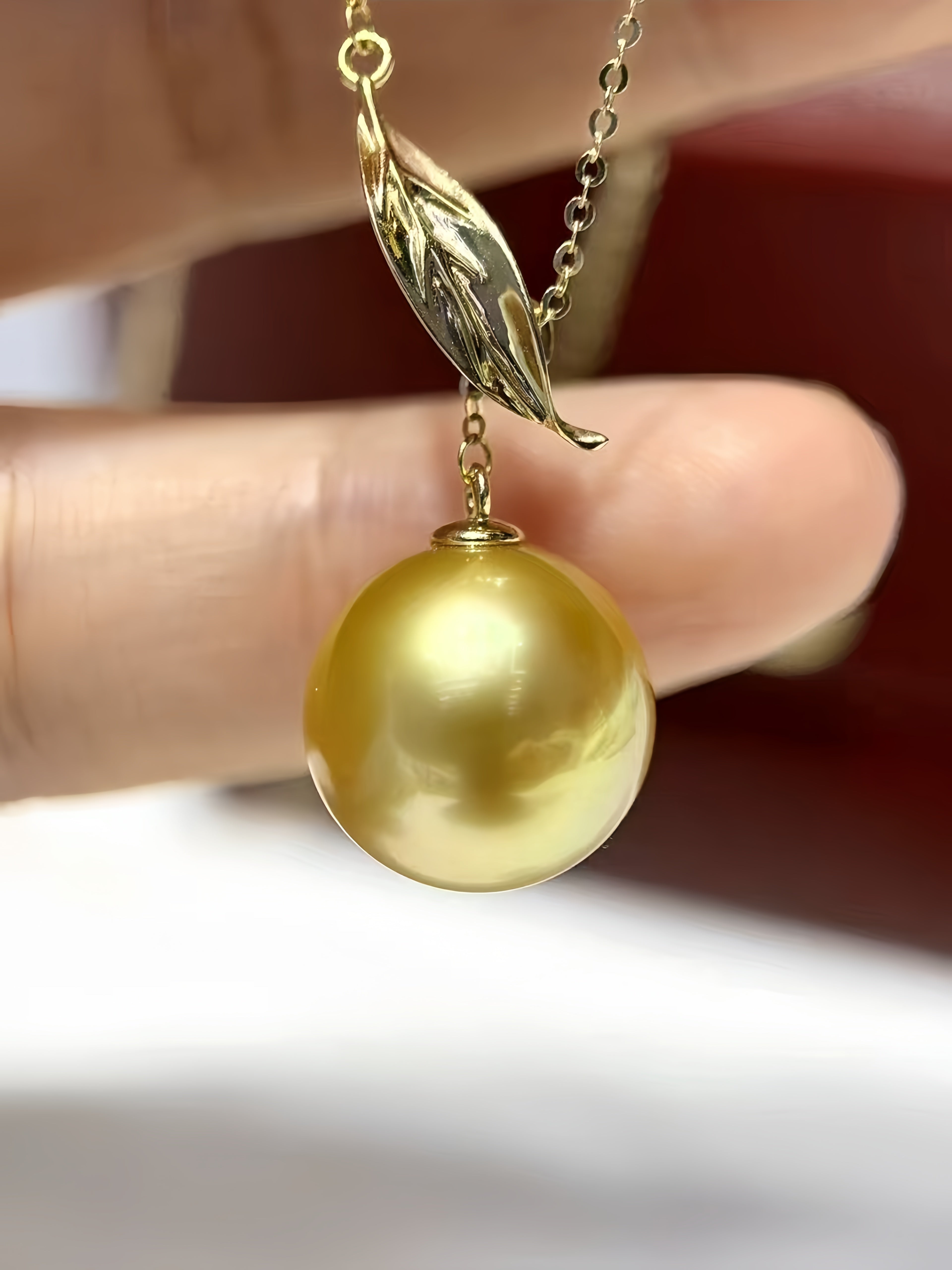 A gold pendant necklace featuring a large, round, lustrous gold pearl suspended from a leaf-shaped pendant on a delicate chain, held by fingers against a blurred background.