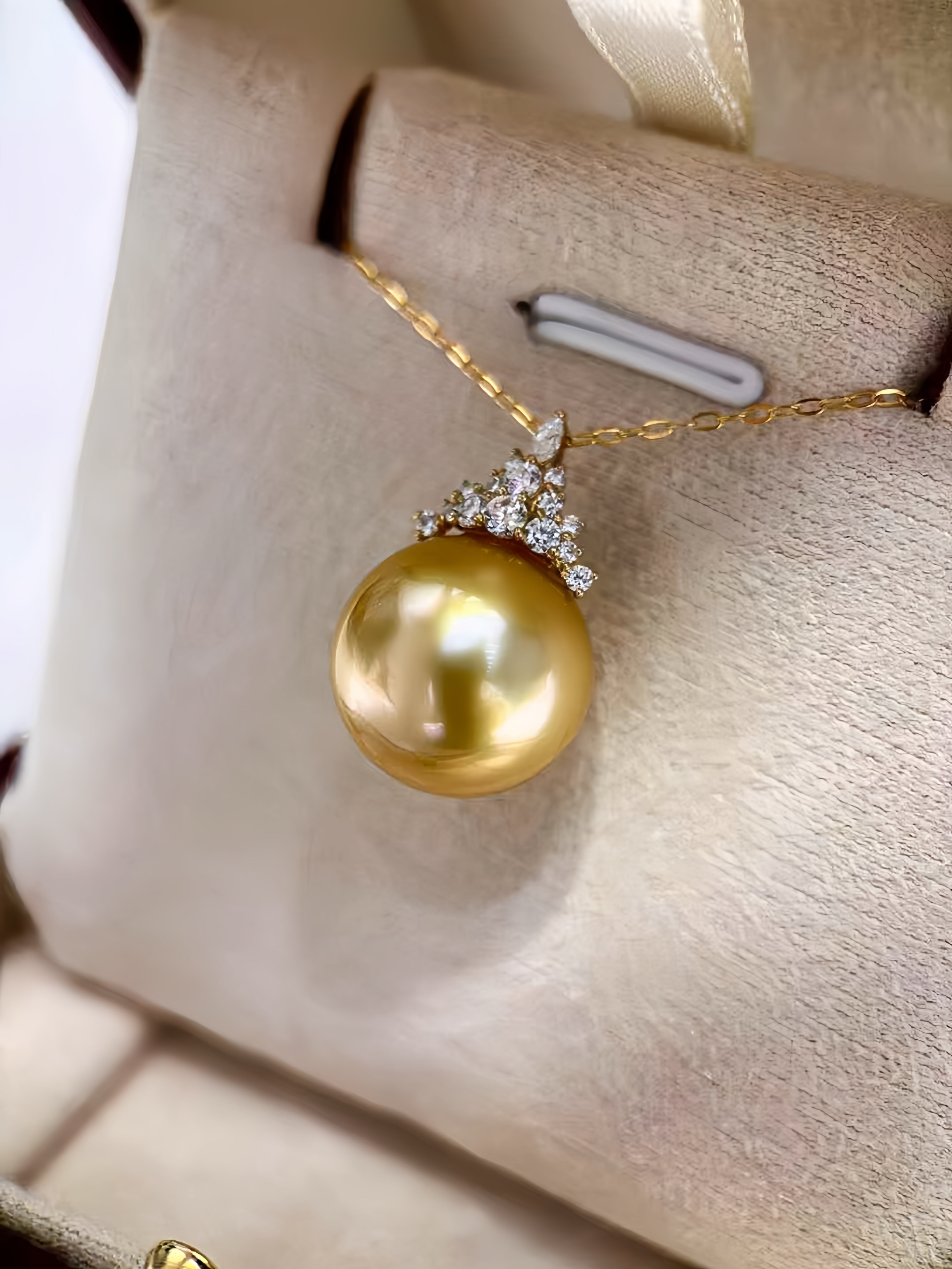 A gold pendant necklace featuring a large, lustrous golden pearl suspended from a delicate gold chain, accented with small clear stones at the top, displayed on a beige jewelry stand.