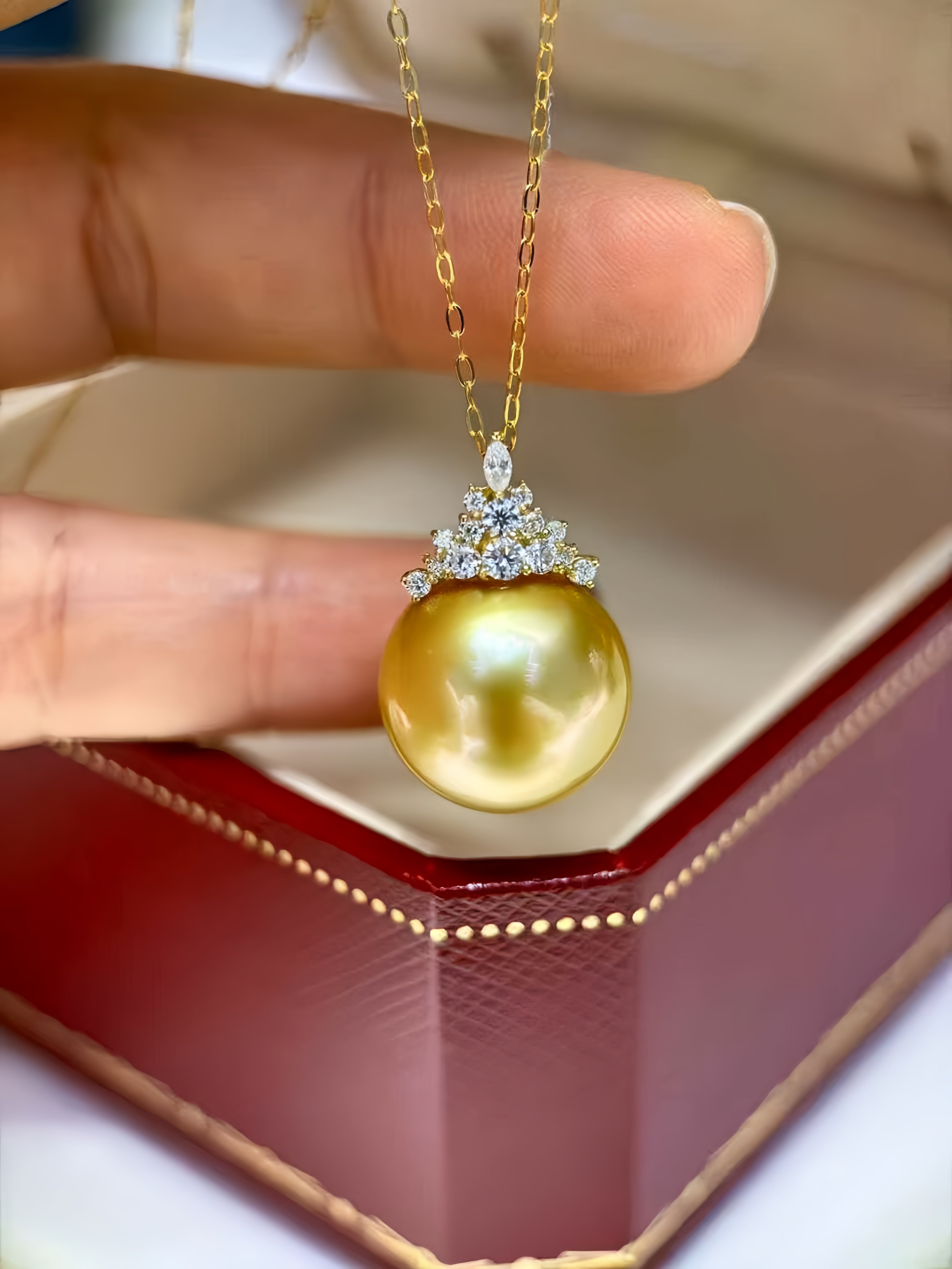 A close-up of a hand holding a gold pendant necklace with a large round yellow-gold pearl and a small clear stone crown setting, positioned over an open dark red jewelry box.