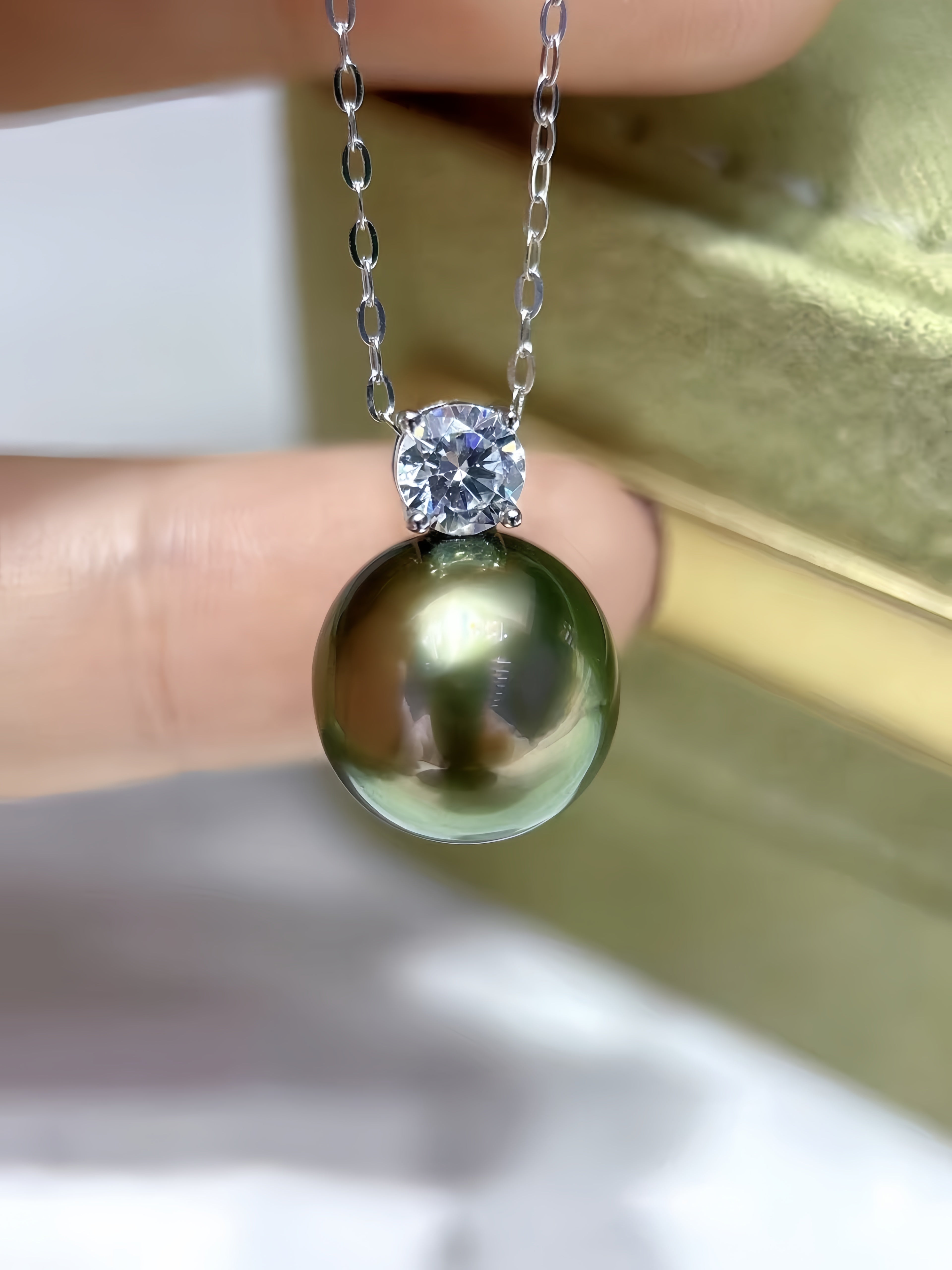 A close-up of a necklace being held between fingers, featuring a large round dark green pearl pendant set with a clear round gemstone on a silver chain.