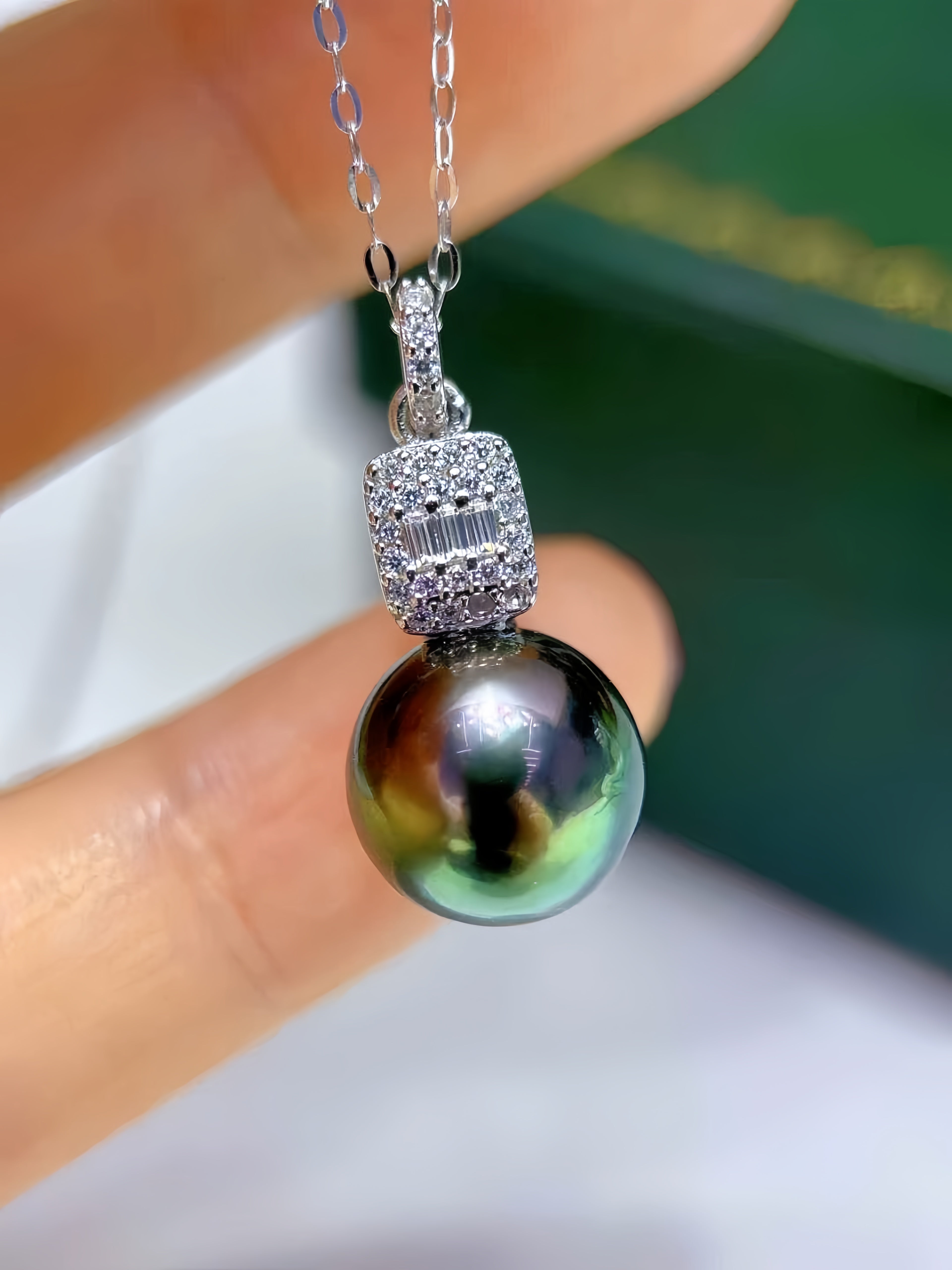 A close-up of a silver pendant necklace held between fingers, featuring a large iridescent green-black pearl and a silver bezel-set pavé top with clear stones.