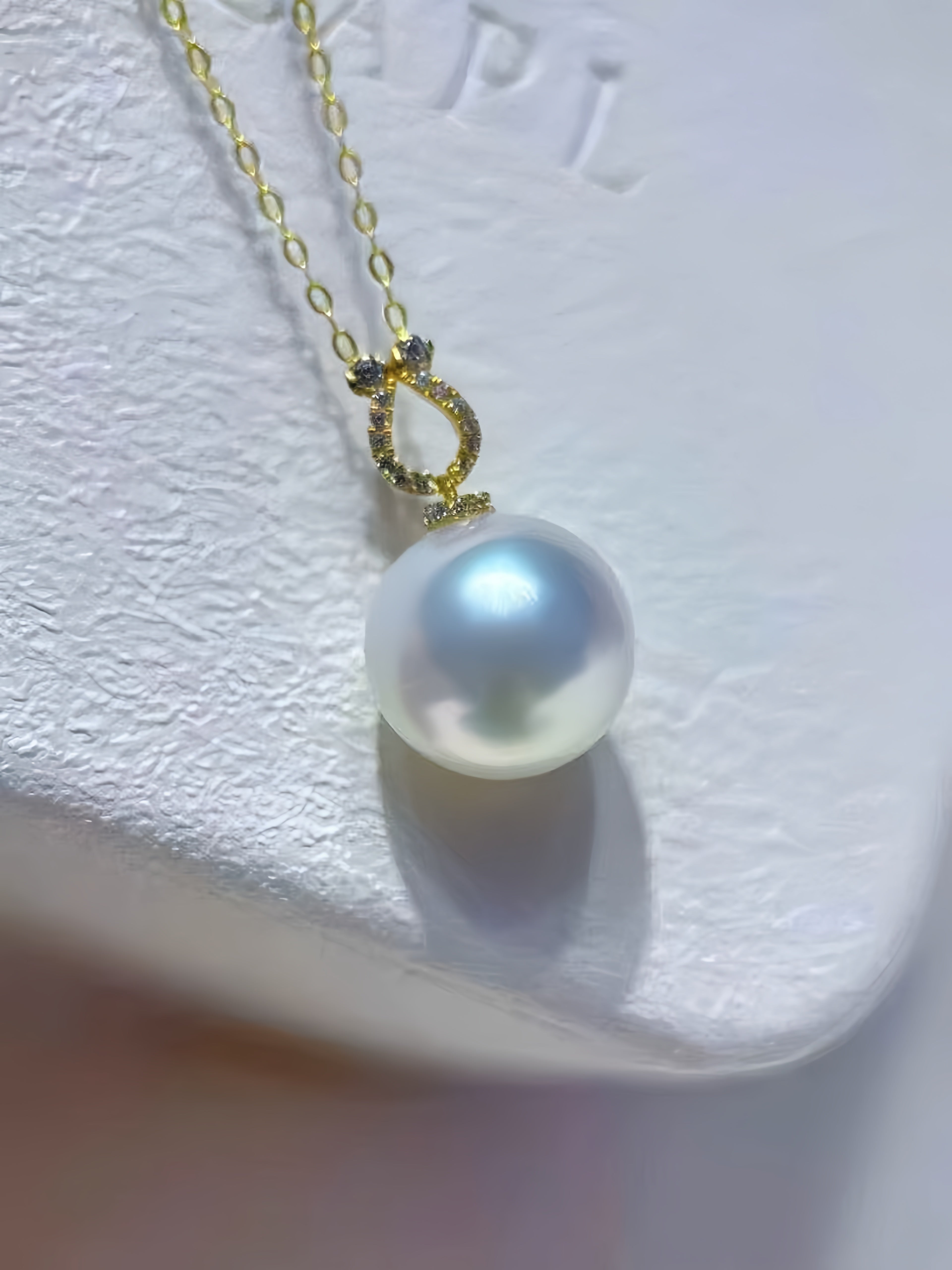 A gold chain necklace with a single lustrous white pearl pendant resting on a textured white surface.