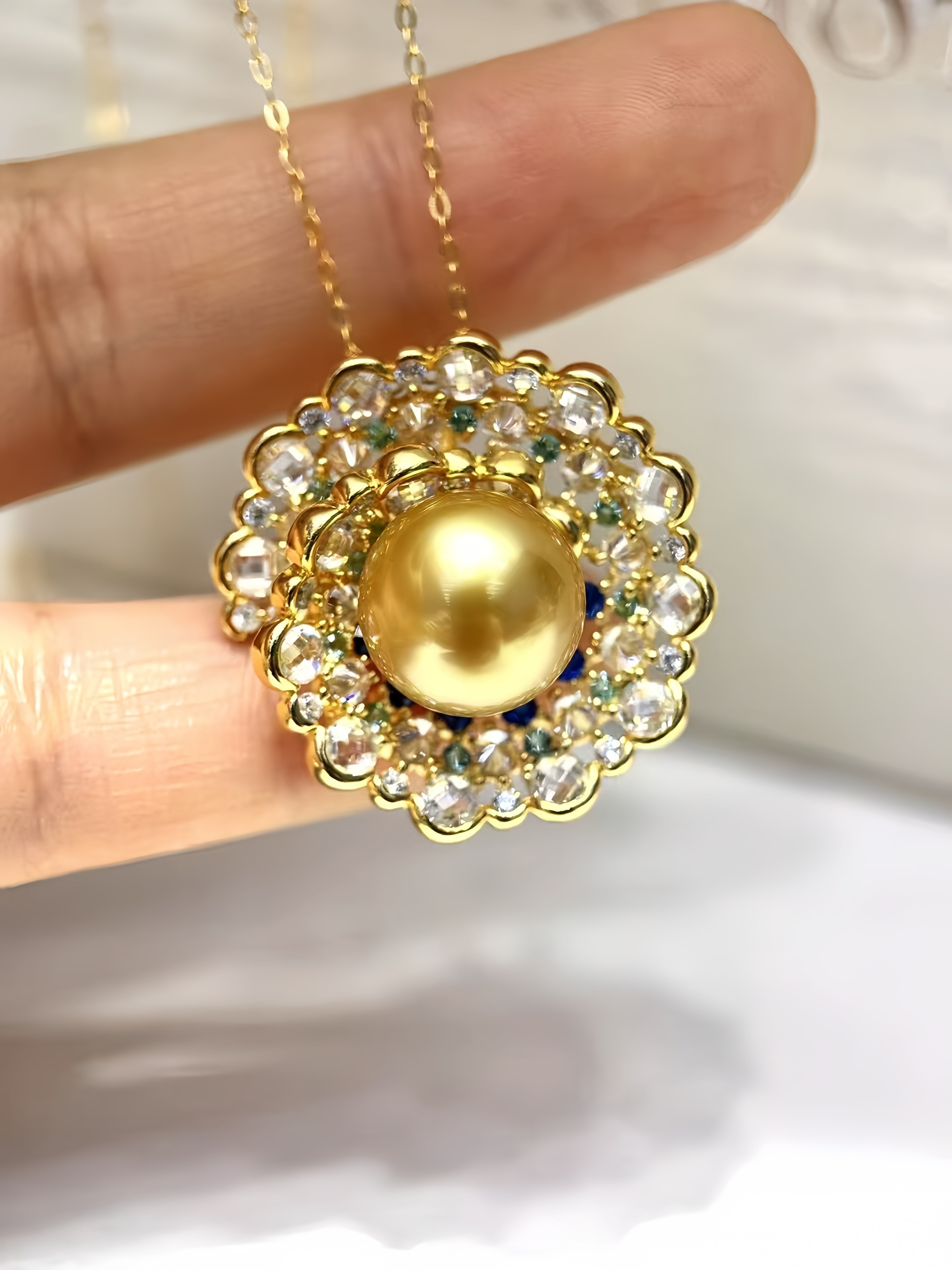 A gold pendant necklace with a large central gold pearl surrounded by clear rhinestones and small green and blue accent stones, held between fingers.