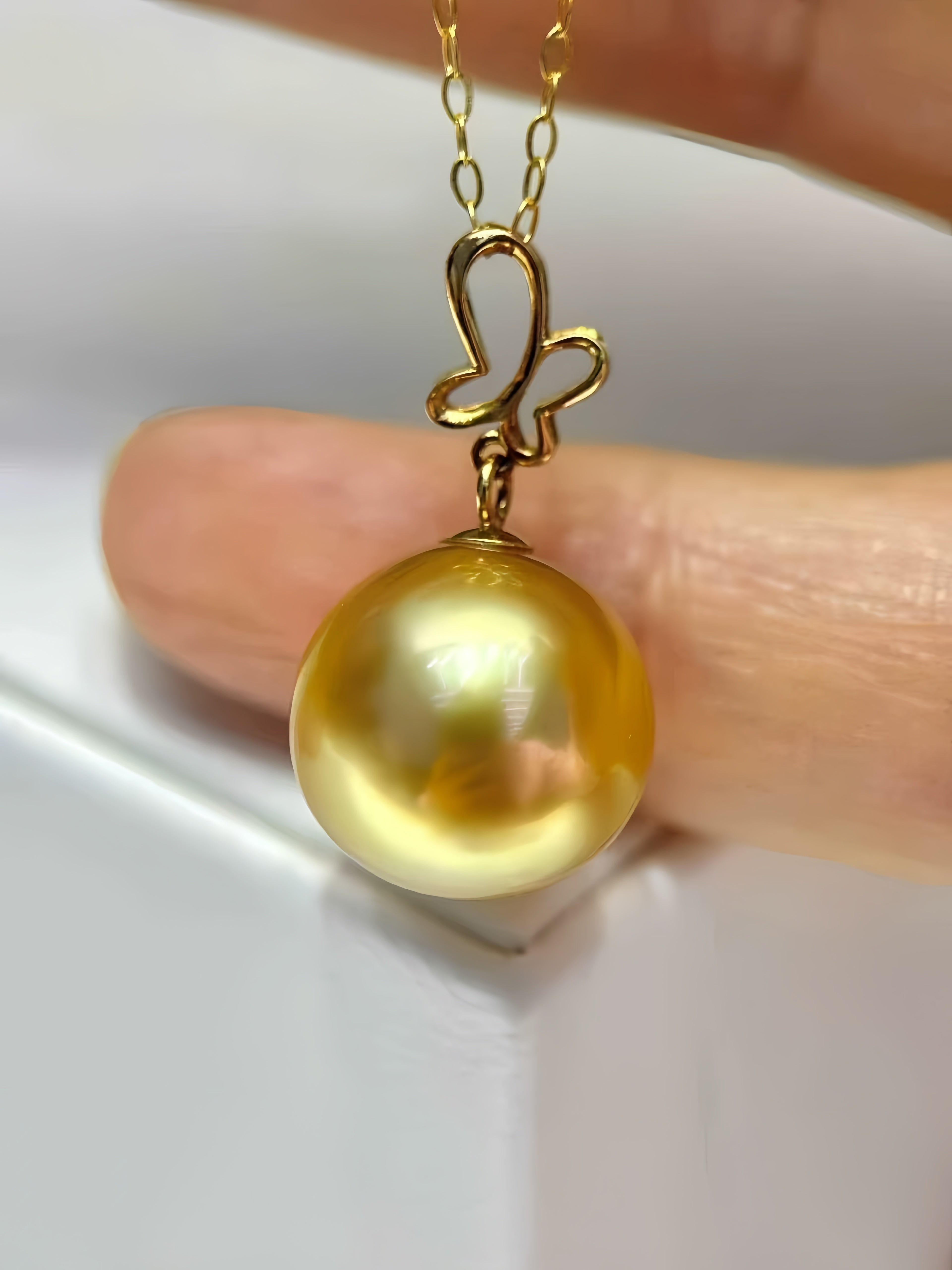 A close-up of a gold pendant necklace featuring a large, lustrous golden pearl and a butterfly-shaped gold bail, held between fingers.