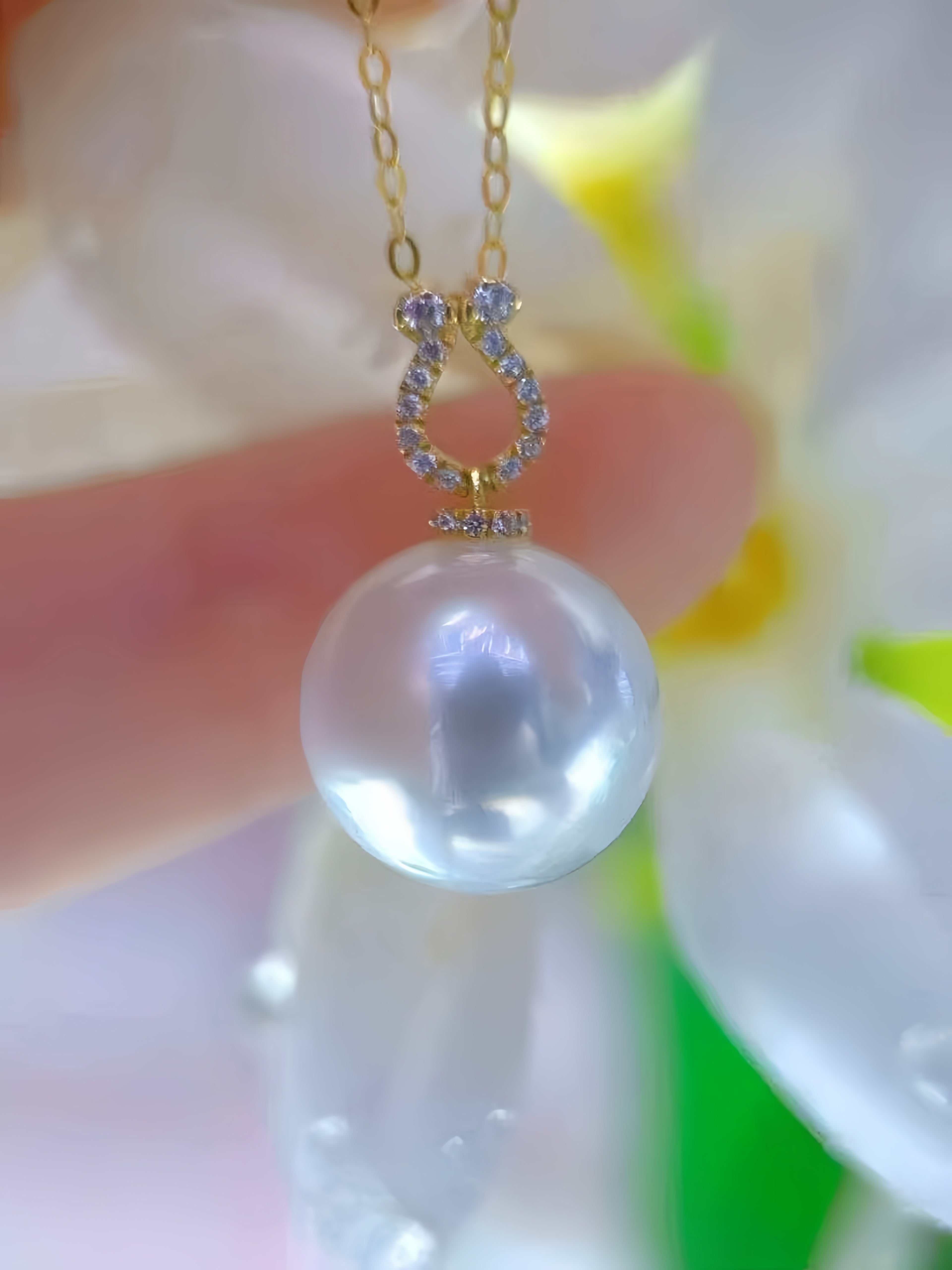 A gold pendant necklace with a large, lustrous white pearl and a horseshoe-shaped setting accented with small clear stones, held near a flower.