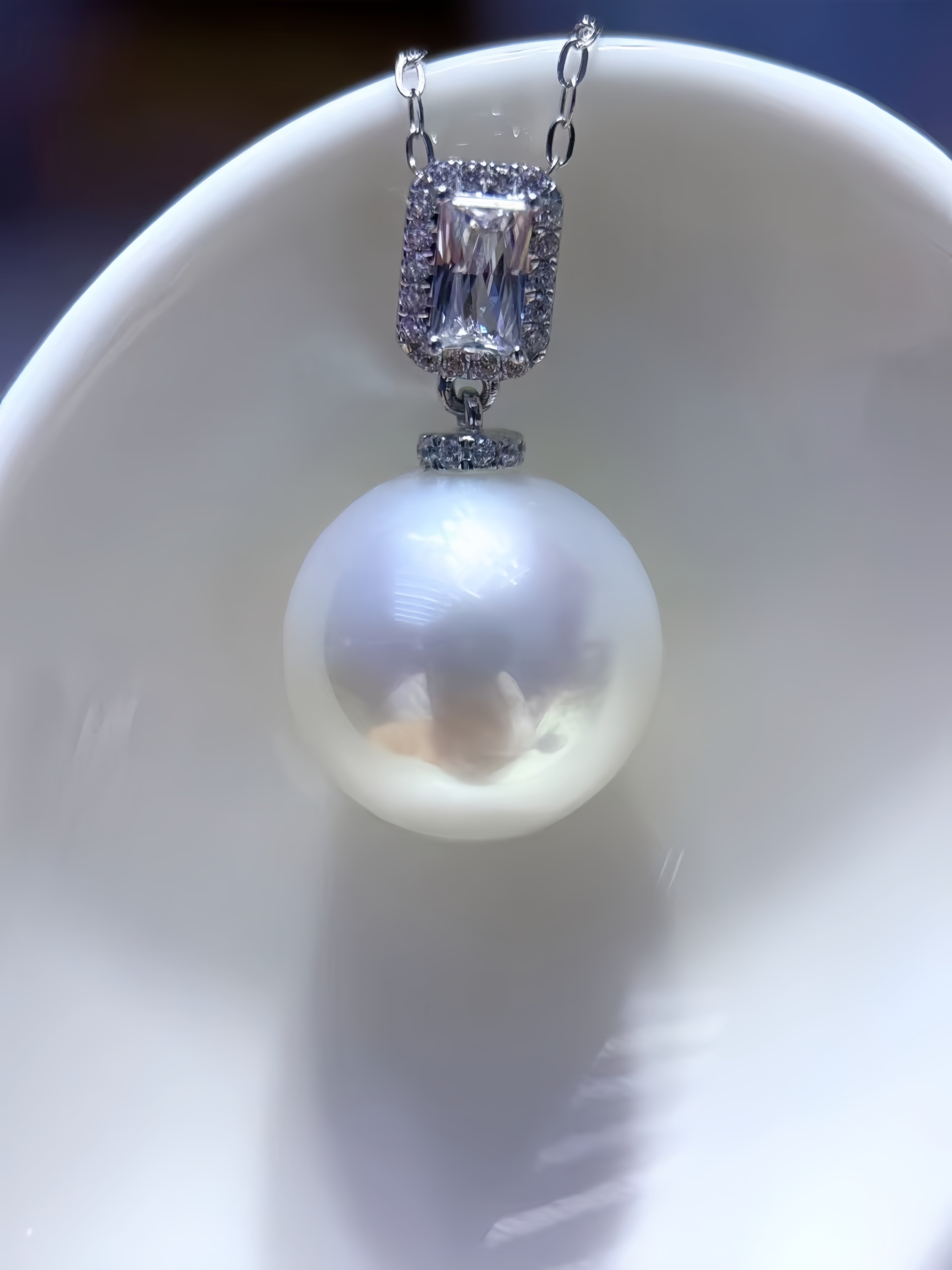 A close-up of a pendant necklace featuring a large, lustrous white pearl suspended below a silver-toned rectangular bezel setting with small clear stones, resting on a white ceramic dish.