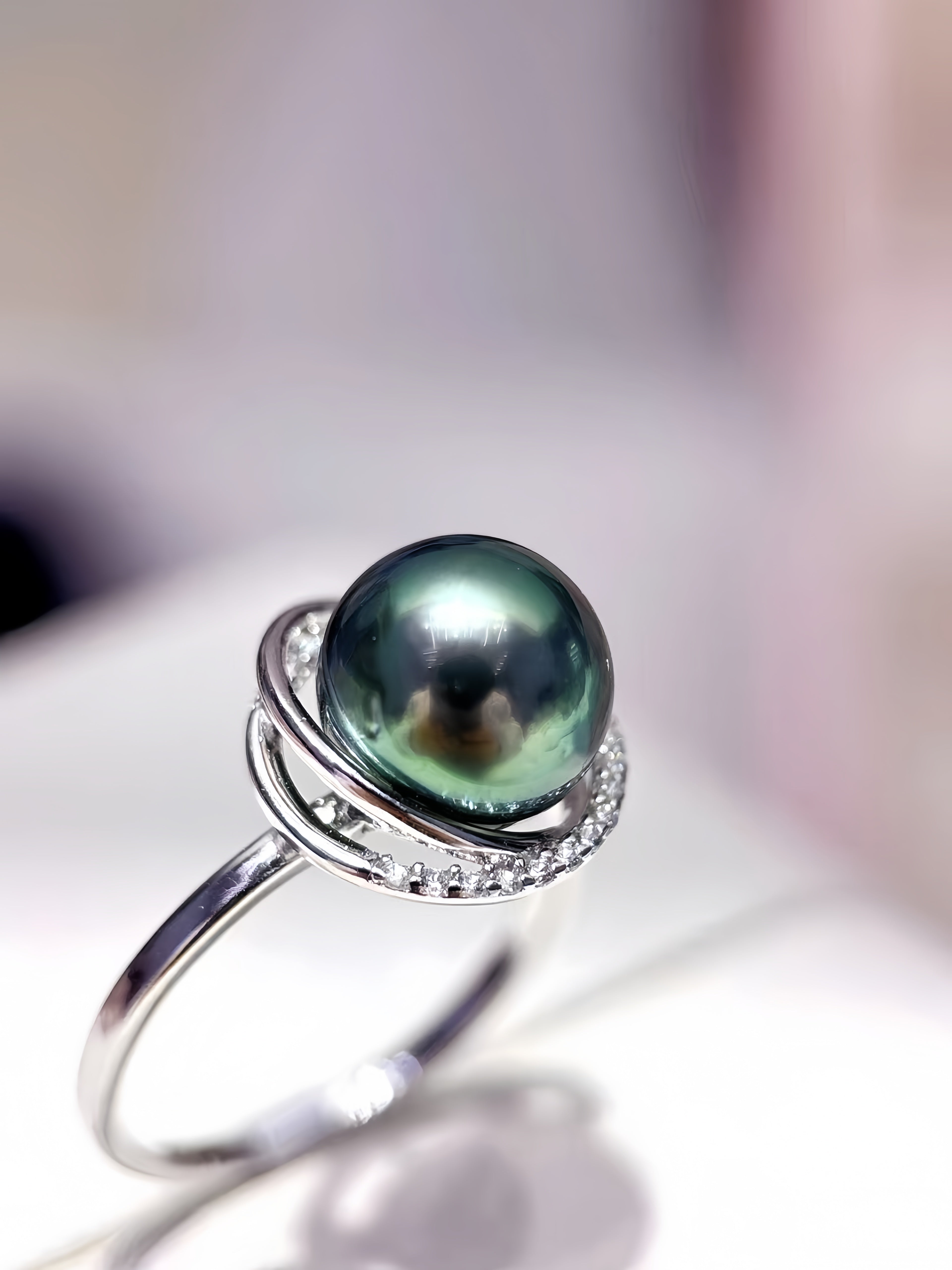 A silver ring featuring a large, round, dark green-black Tahitian pearl surrounded by small clear stones and a swirling silver setting, displayed on a soft-focus white surface.
