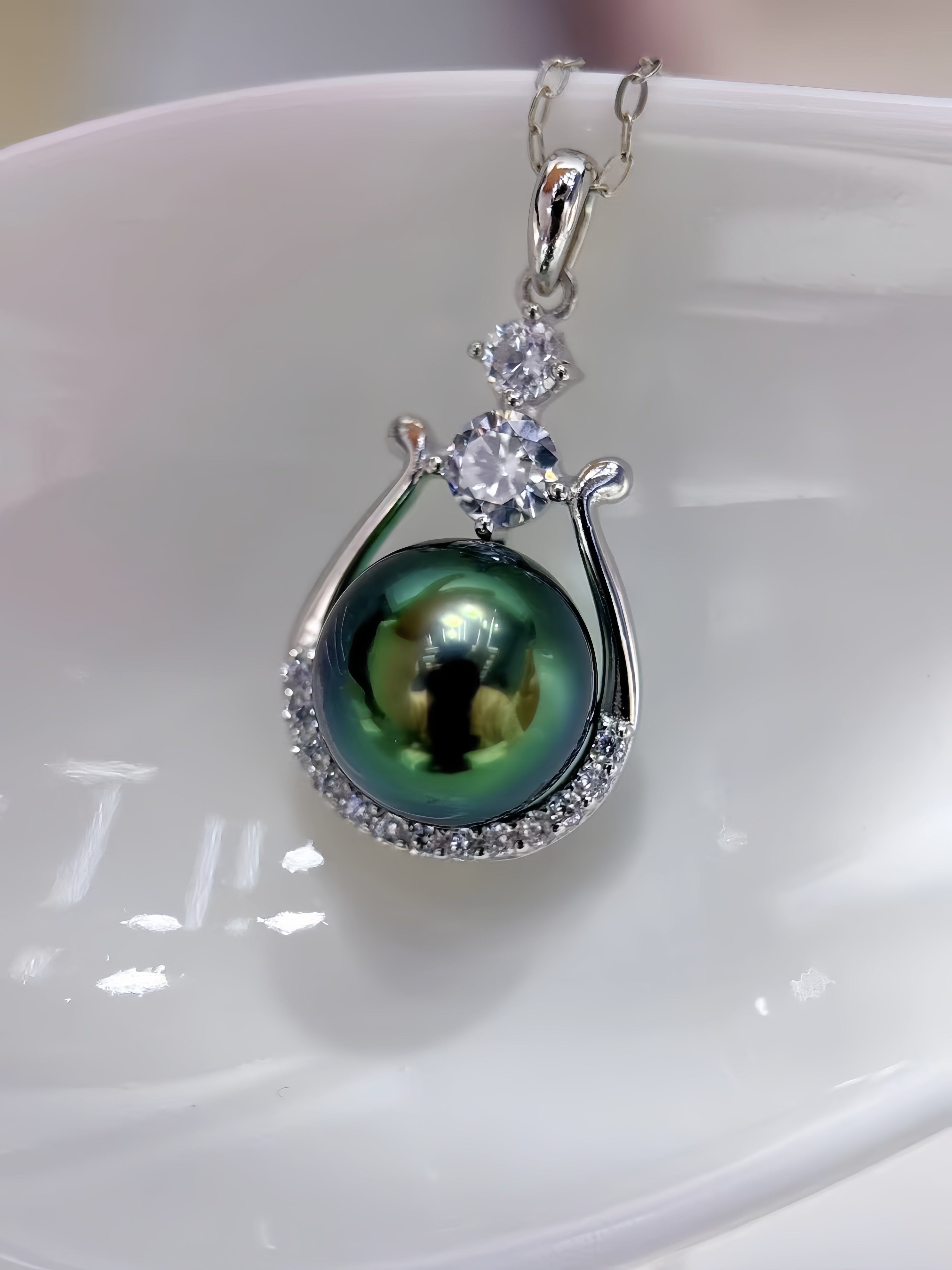 A silver pendant necklace featuring a large, round, dark green-black Tahitian pearl surrounded by a halo of small clear stones, with a three-stone cluster at the top, resting on a glossy white ceramic dish.