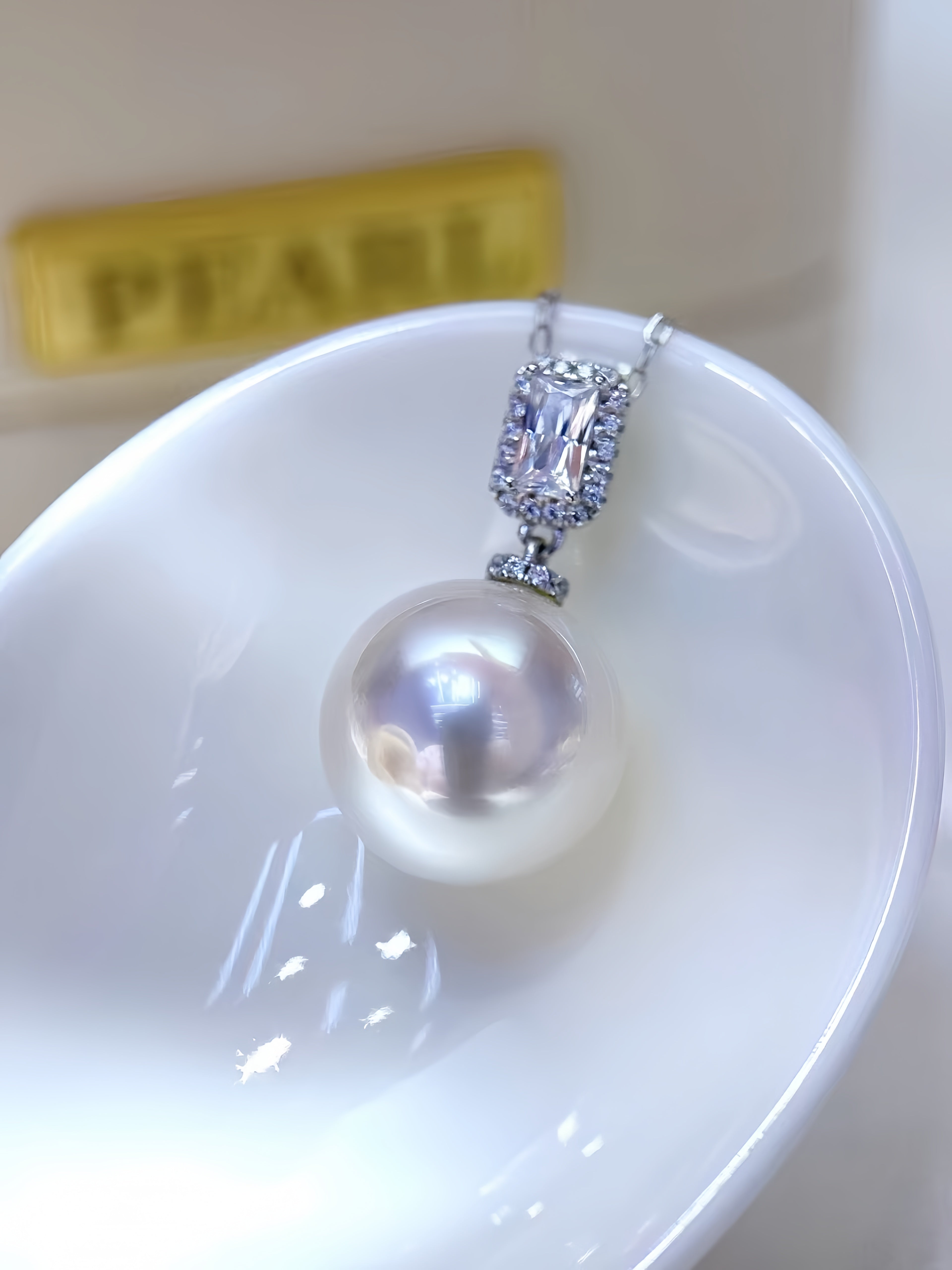 A pearl pendant necklace with a silver-toned halo setting, resting on a white ceramic dish with a blurred gold 'PEARL' sign in the background.