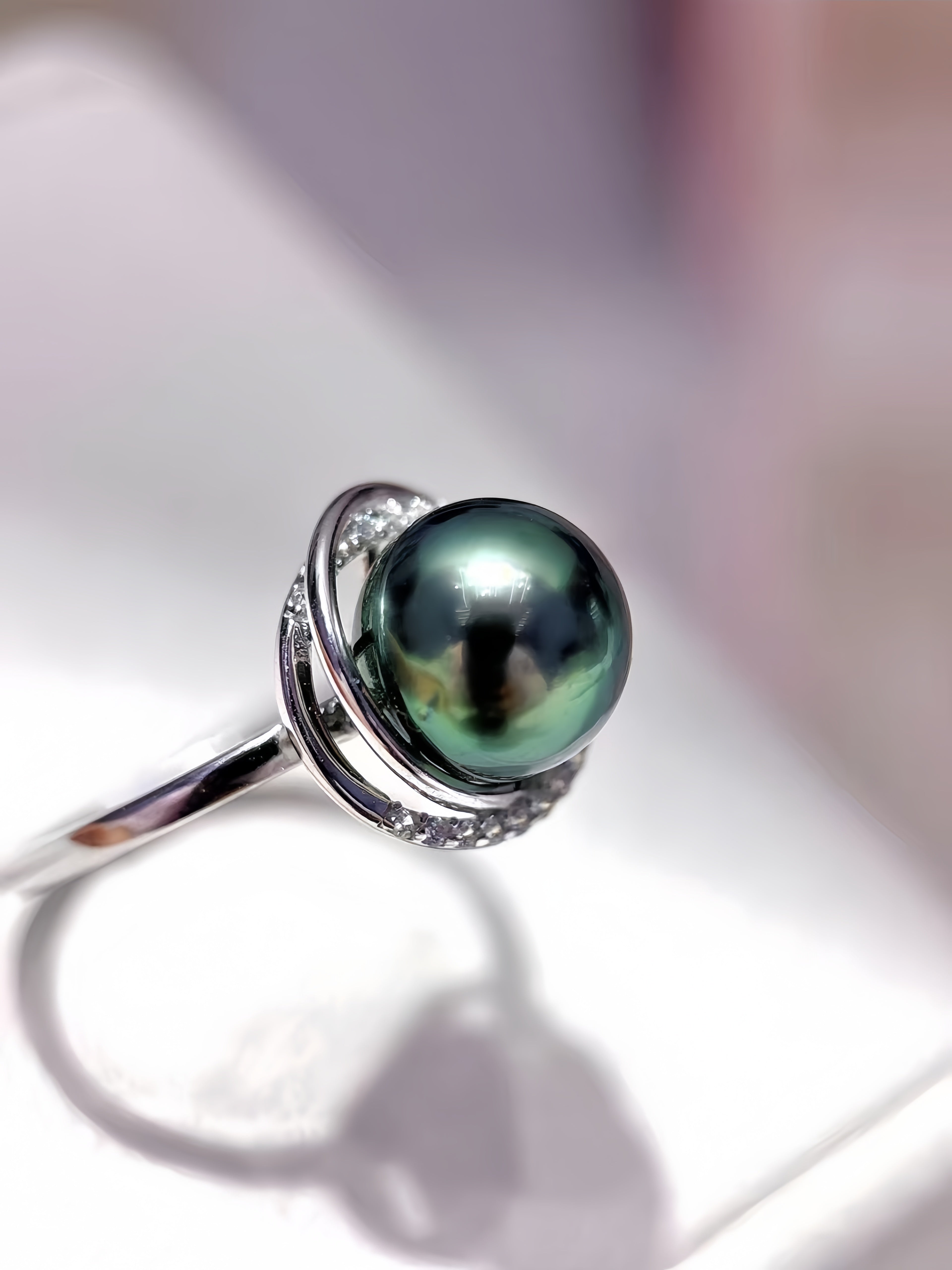 A sterling silver ring featuring a large, round, iridescent green-black Tahitian pearl set in a double-ring halo with small accent stones, shown on a soft-focus white background.