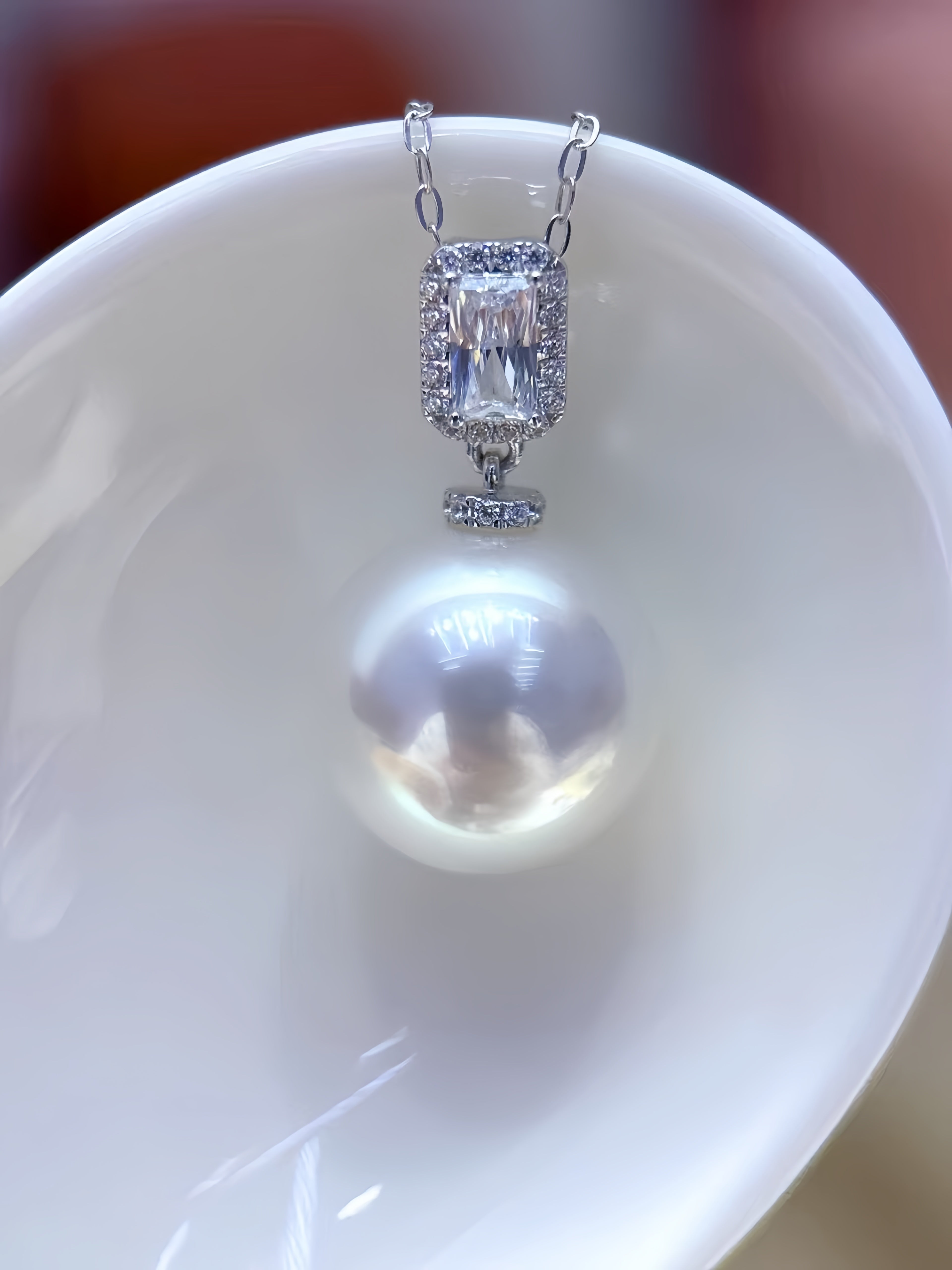 A close-up of a silver pendant necklace featuring a large rectangular clear gemstone surrounded by small clear stones, with a large white pearl hanging below, displayed against a glossy white curved surface.