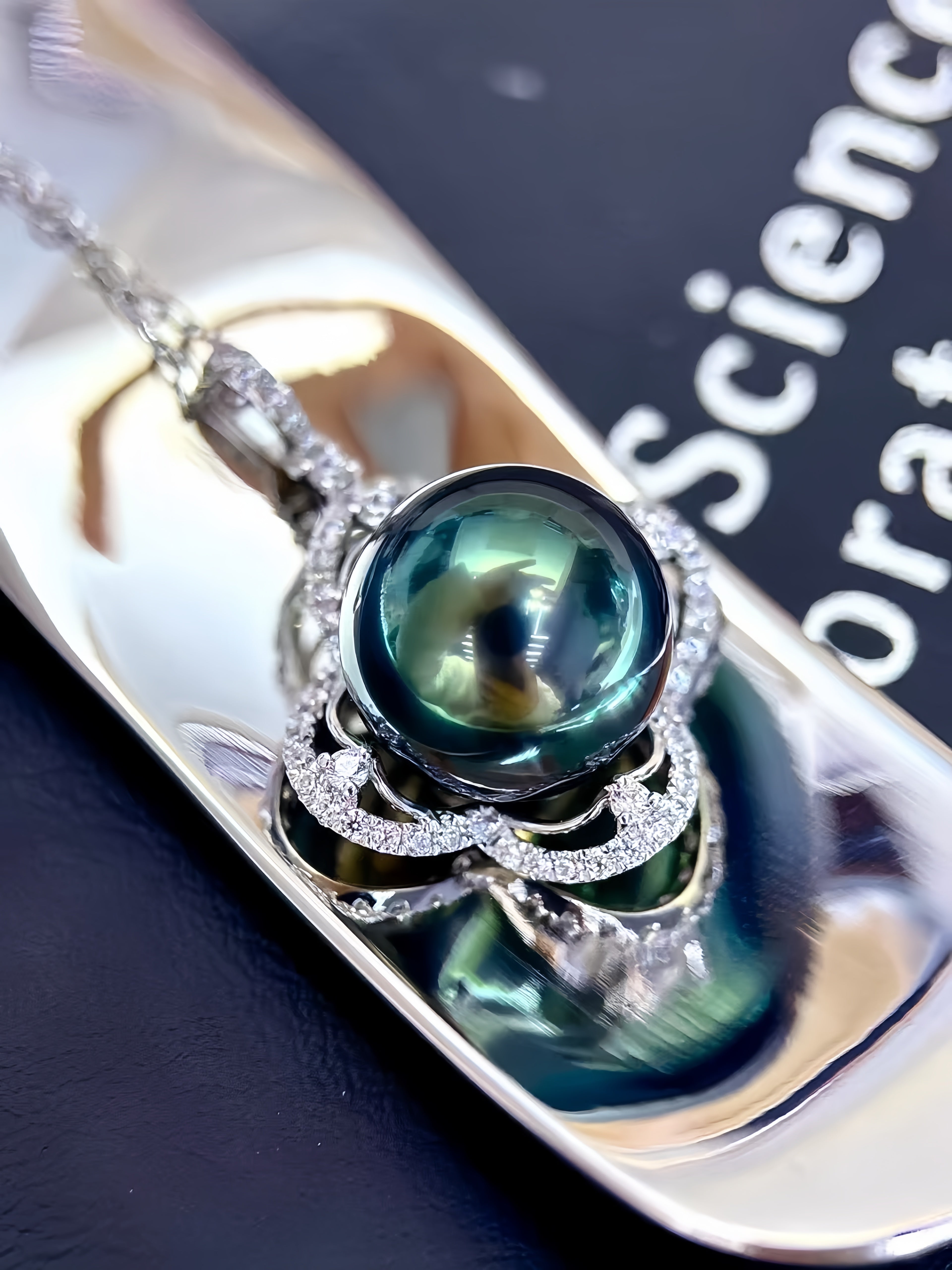 A close-up of a silver pendant necklace featuring a large, round, dark green teardrop-shaped pearl surrounded by a halo of small clear stones, resting on a polished silver tray.