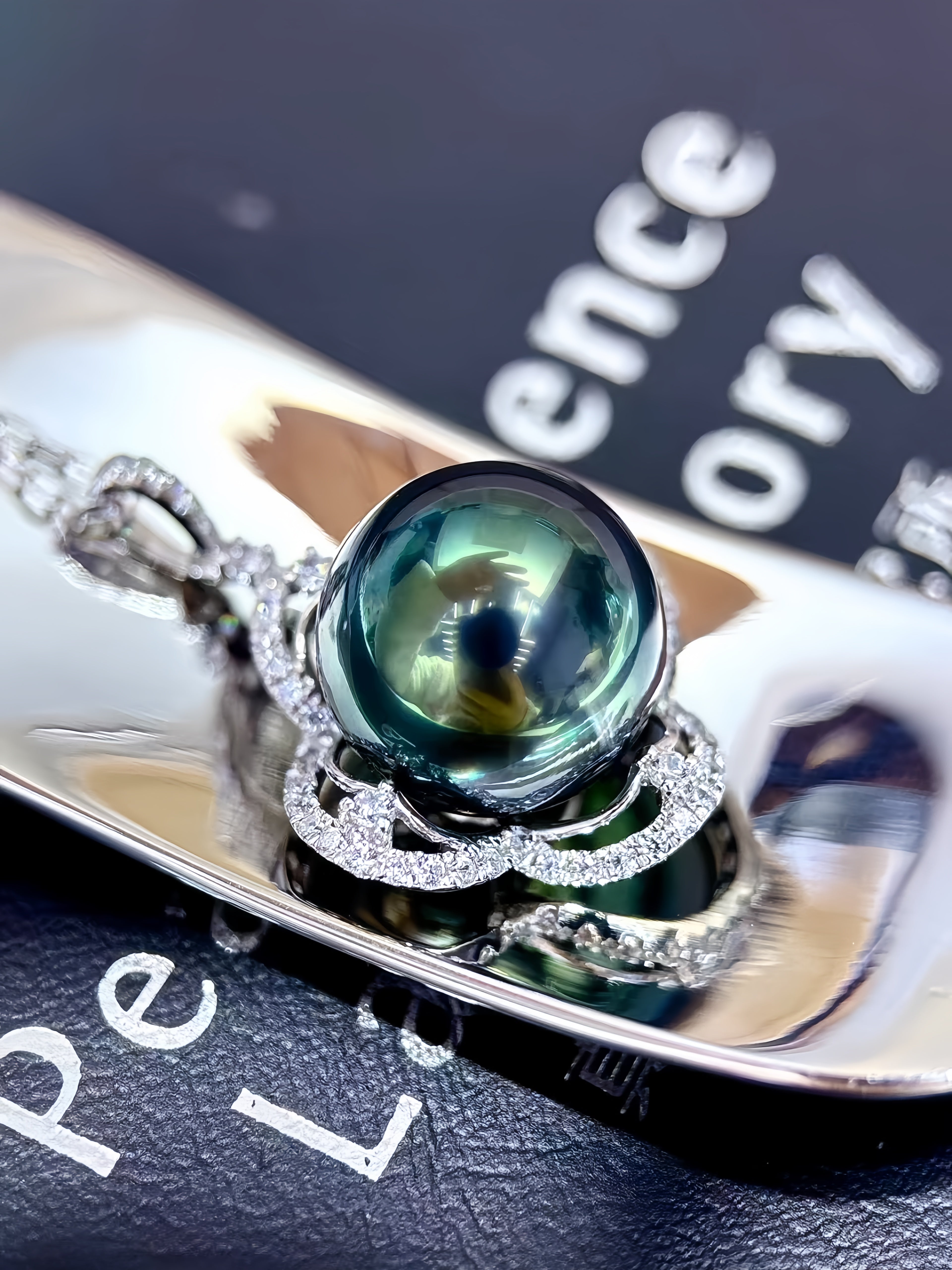 A close-up of a silver pendant necklace featuring a large, round, dark green teardrop-shaped pearl or gemstone, surrounded by intricate silver filigree and small clear stones, resting on a reflective silver tray.