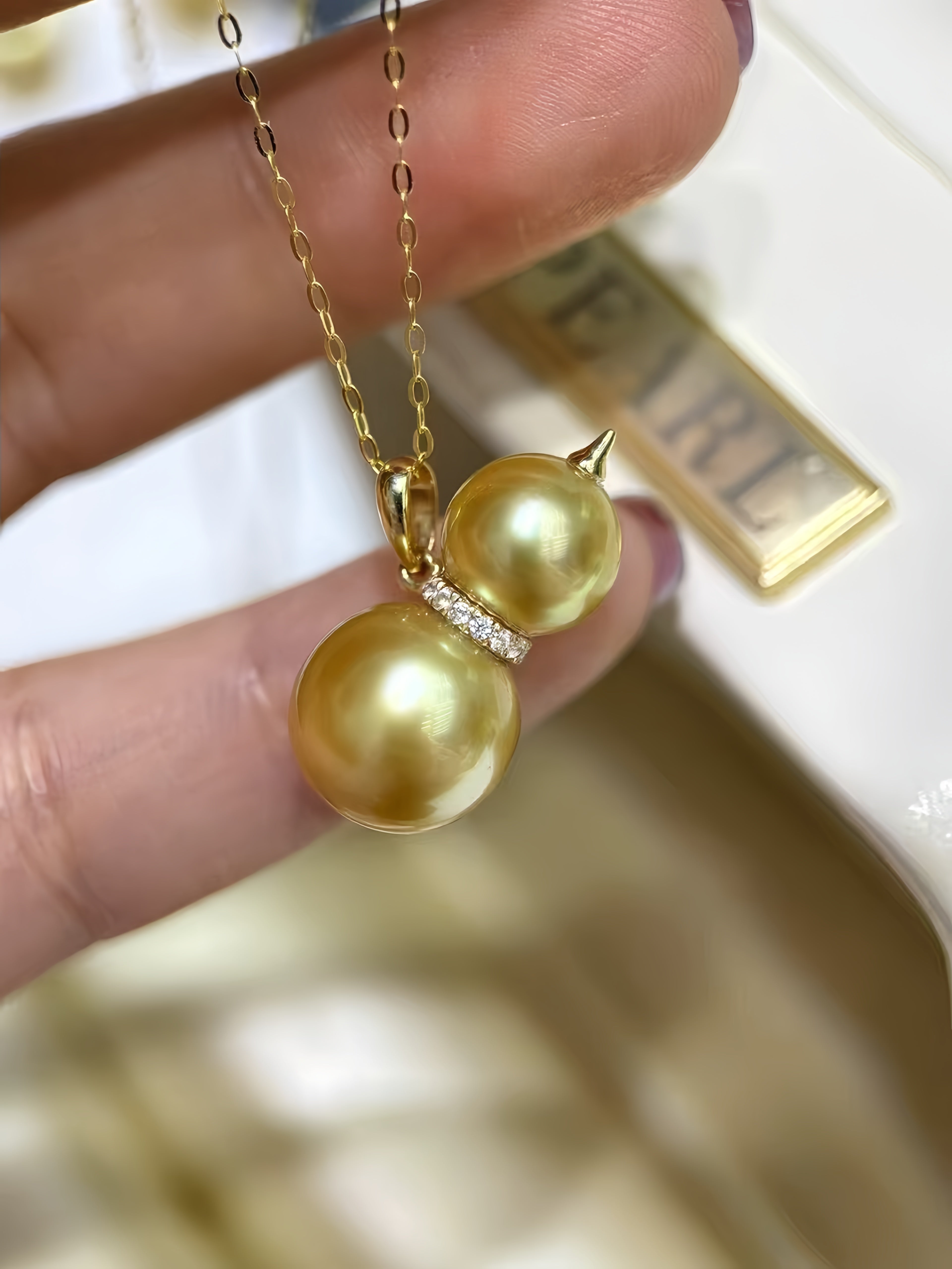 A close-up of a hand holding a gold pendant necklace with two large gold pearls stacked vertically, separated by a small row of clear stones, against a softly blurred jewelry box background.