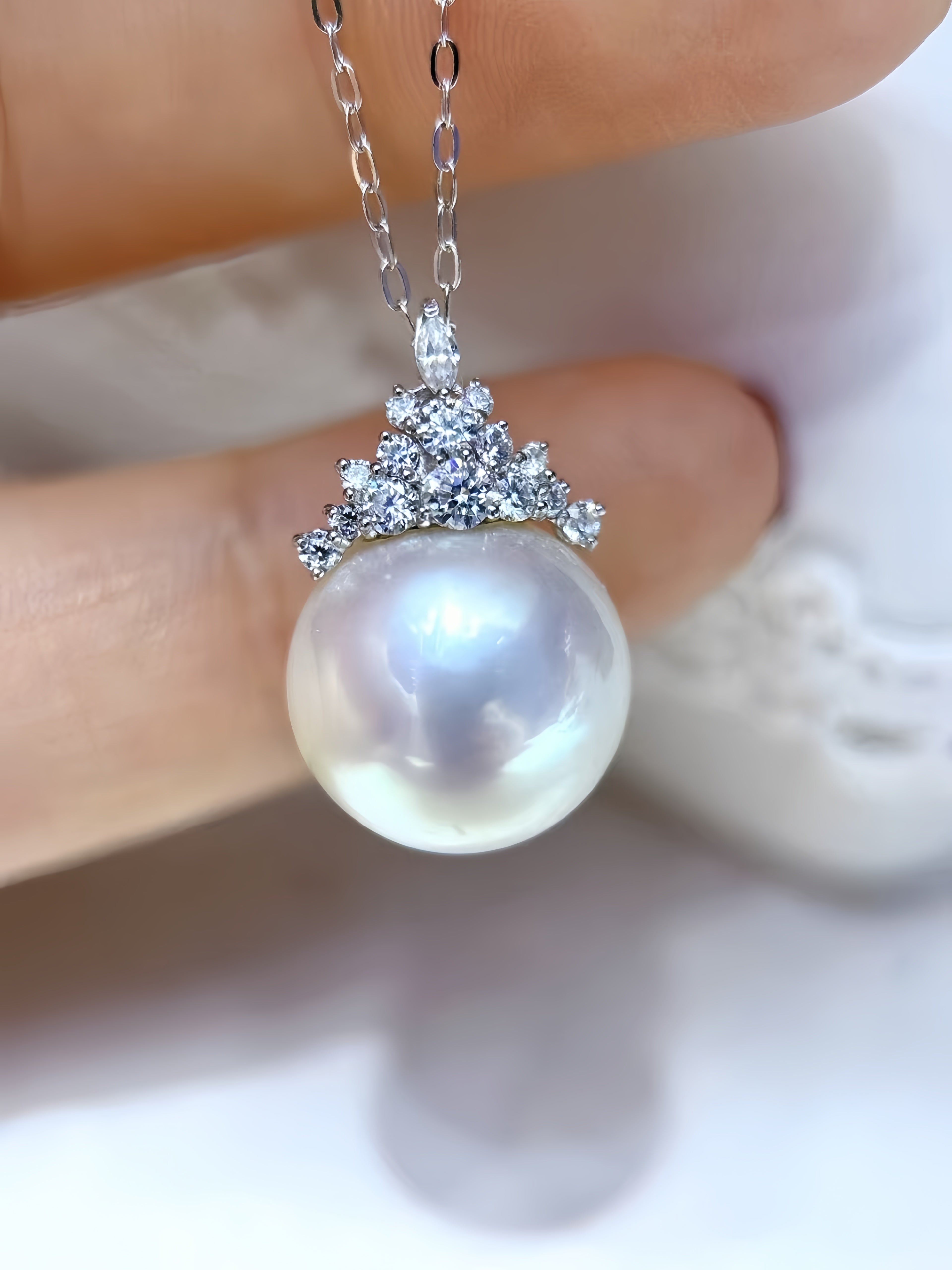 A close-up of a pearl pendant necklace held between fingers. The pendant features a large, lustrous white pearl suspended below a silver-toned crown-like setting adorned with small clear stones, on a delicate silver chain.