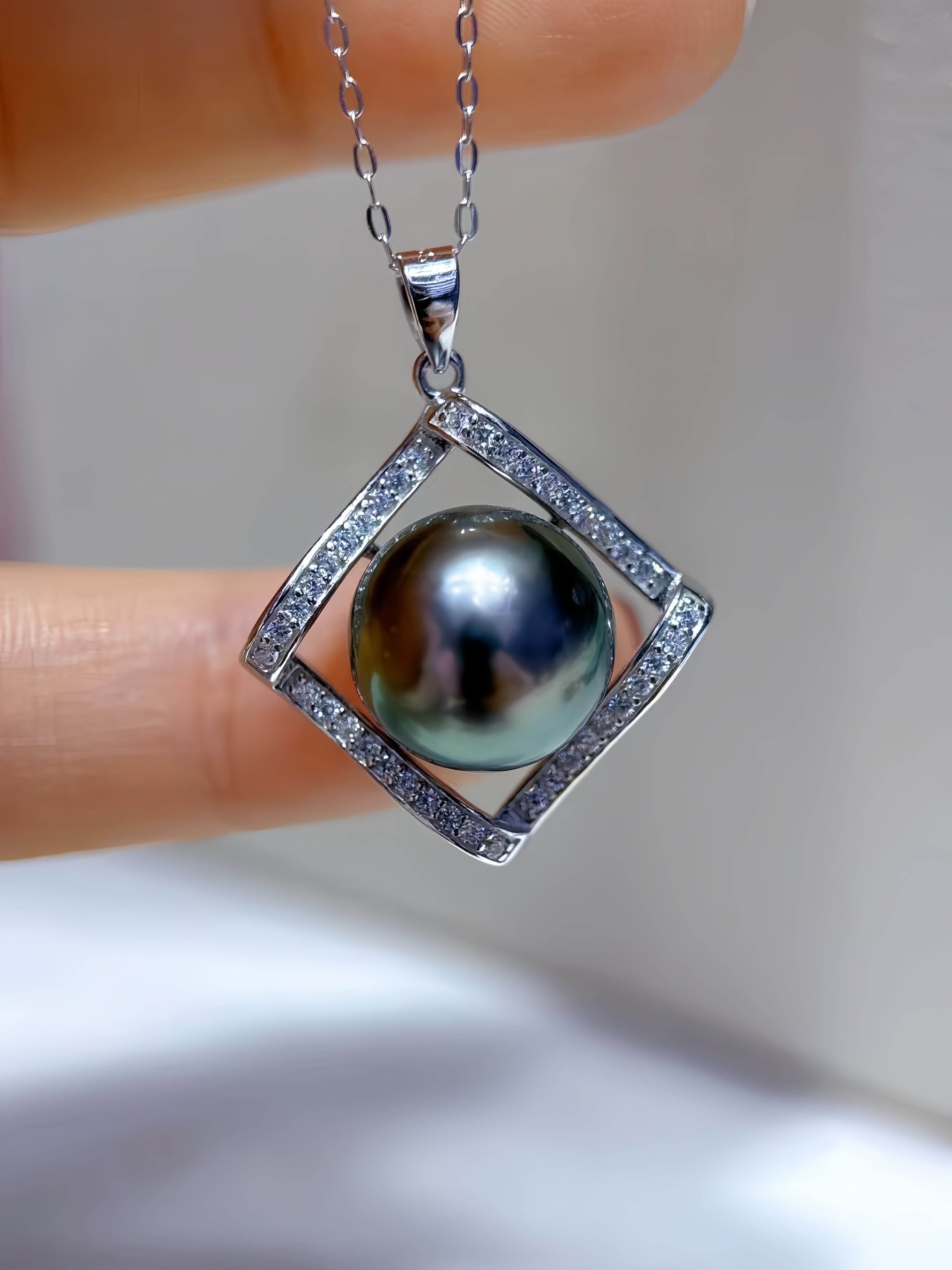 A close-up of a silver pendant necklace held between fingers. The pendant features a large, round, iridescent dark pearl set within a square diamond-shaped frame paved with small clear stones, suspended from a silver chain.