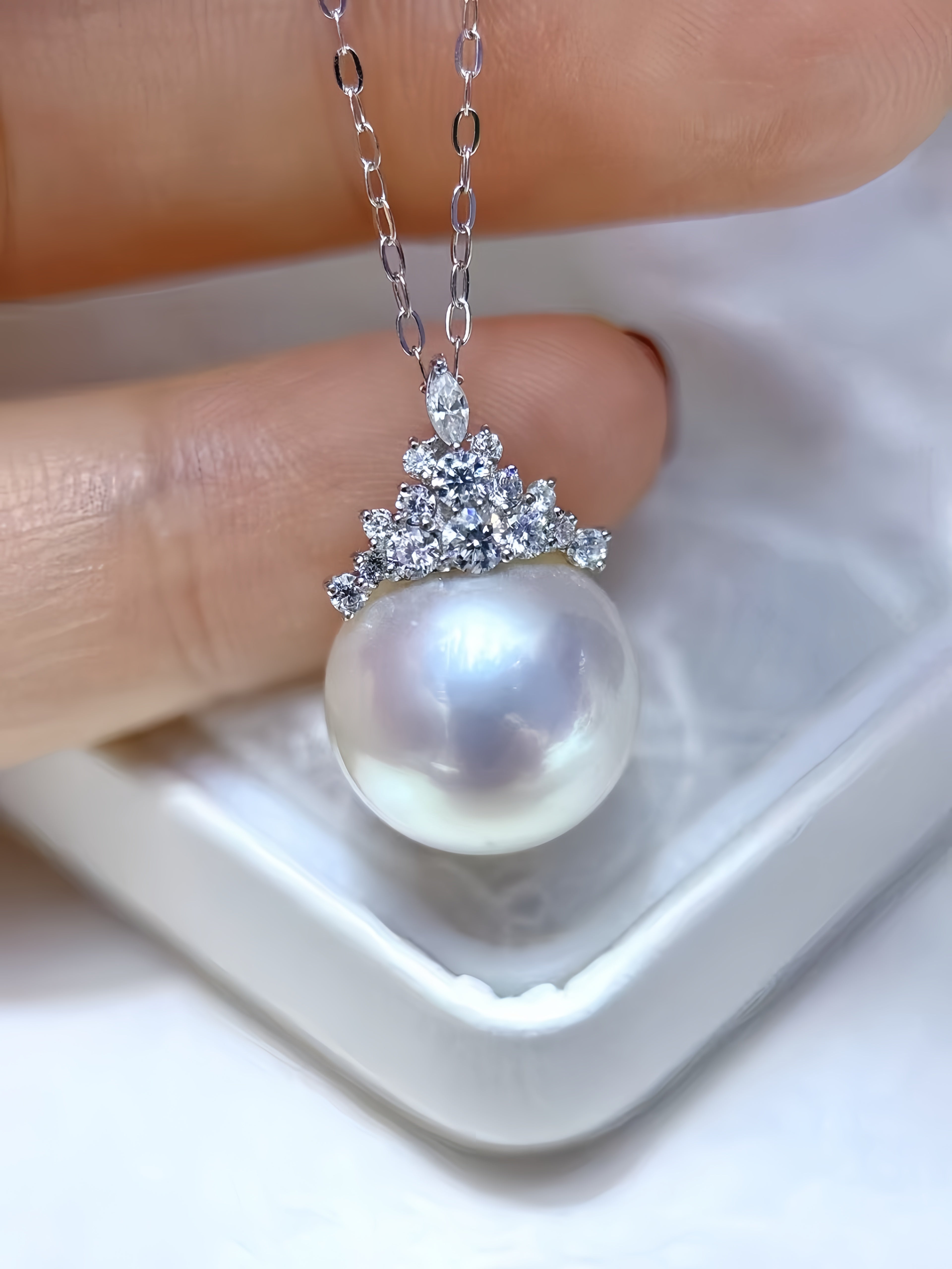 A close-up of a silver pendant necklace with a large lustrous white pearl and a small tiara-like cluster of clear stones, held between fingers above a white jewelry box.