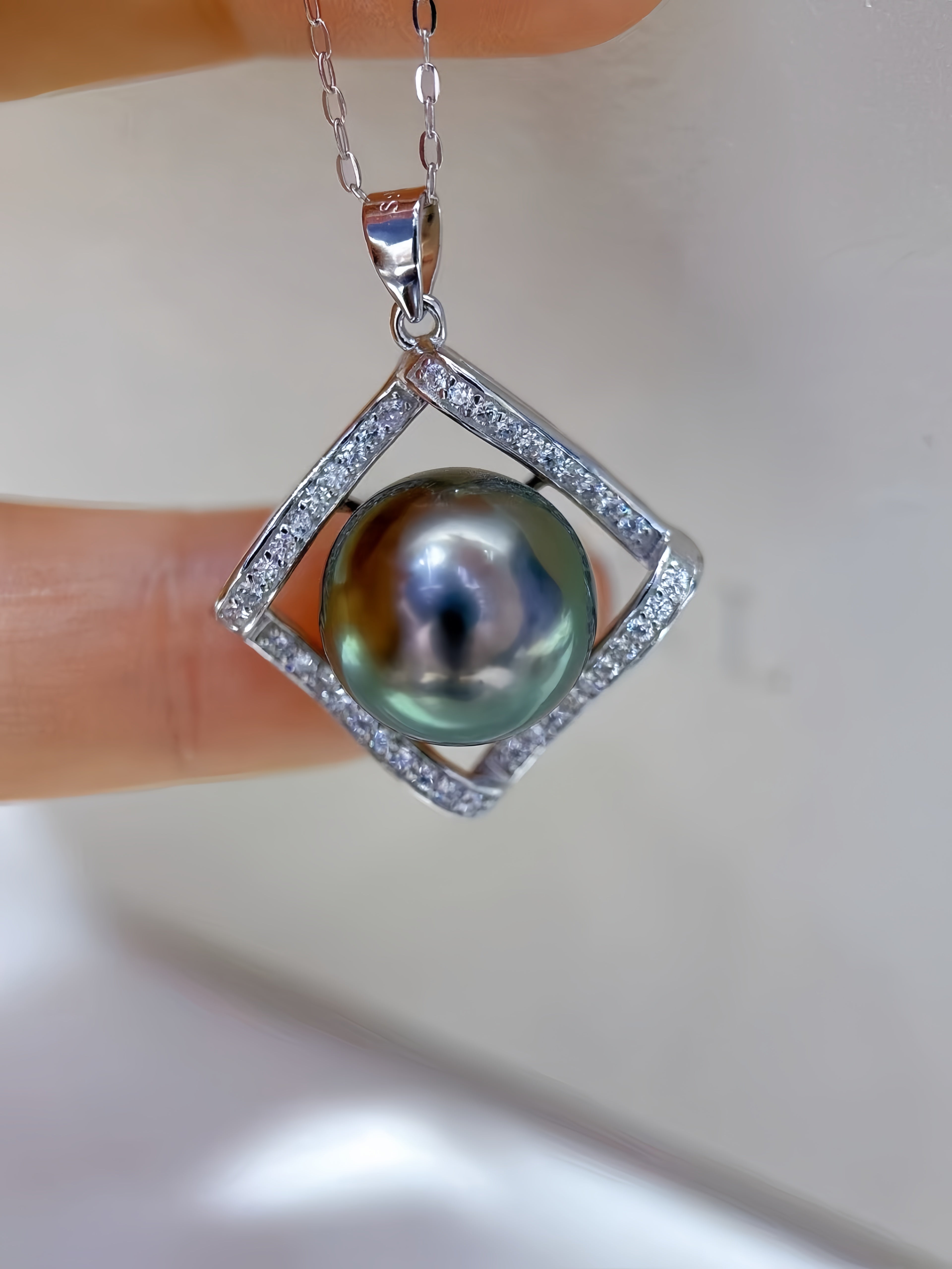 A close-up of a pendant necklace held between fingers. The pendant features a round iridescent dark green pearl set within a silver diamond-shaped frame paved with small clear stones, with a silver chain visible at the top.