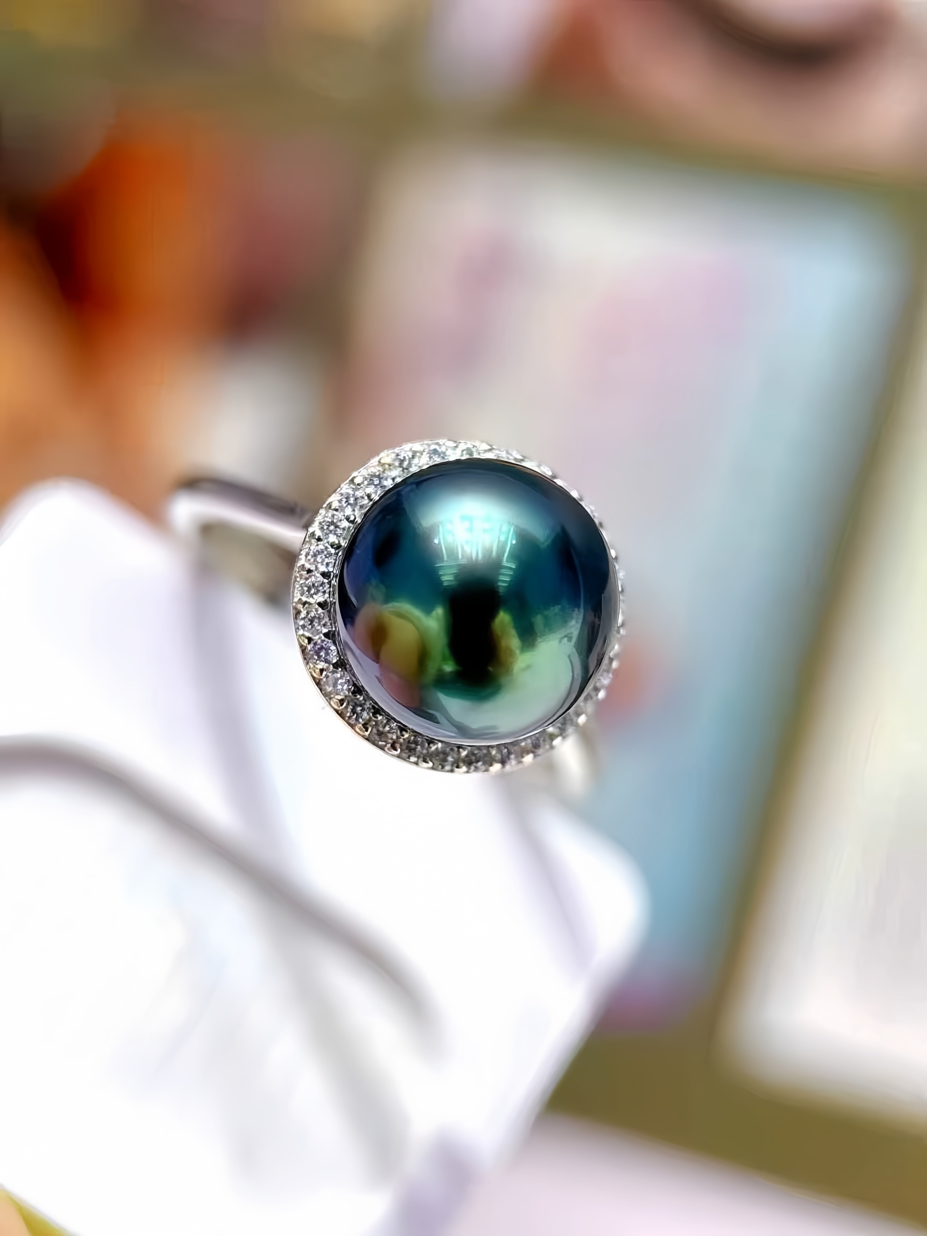 A close-up shot of a silver ring featuring a round, iridescent dark green pearl surrounded by a halo of small clear stones, resting on a white display stand.