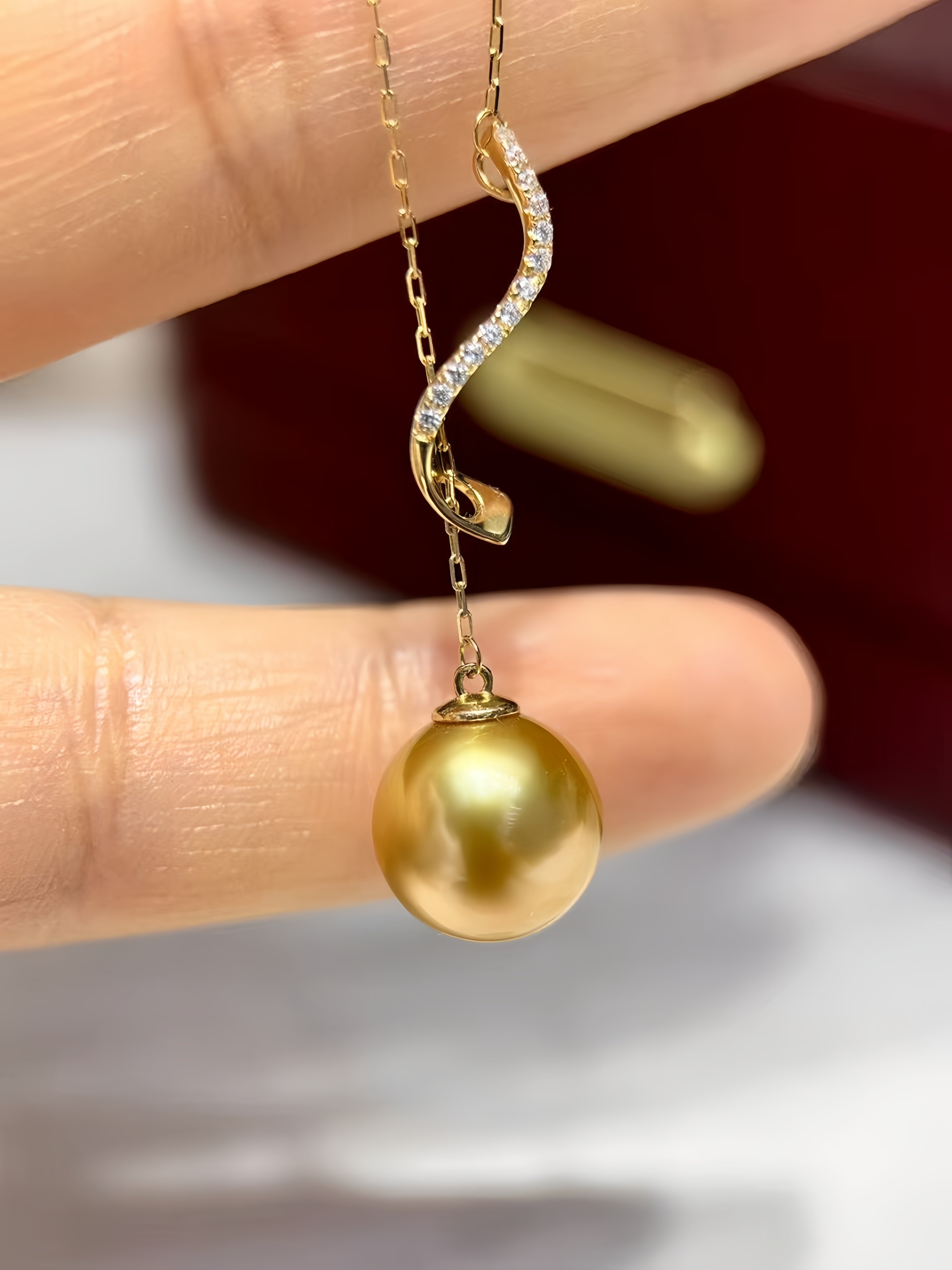 A gold pendant necklace with a curved chain featuring a row of small clear stones and a large round gold pearl, held between fingers against a blurred dark red and white background.