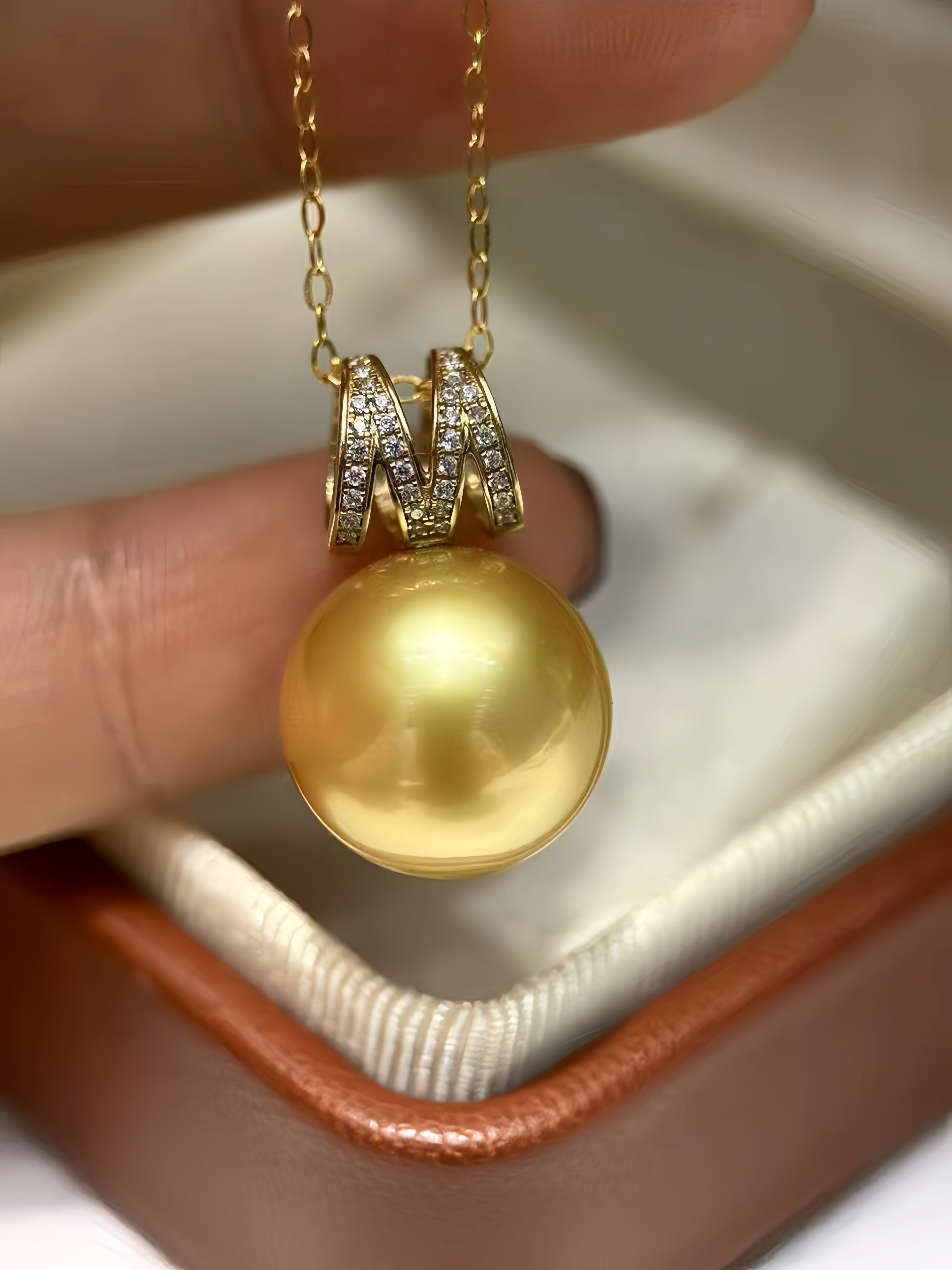 A close-up of a gold pendant necklace with a large round yellow pearl and a diamond-accented bail, held between fingers above an open jewelry box.