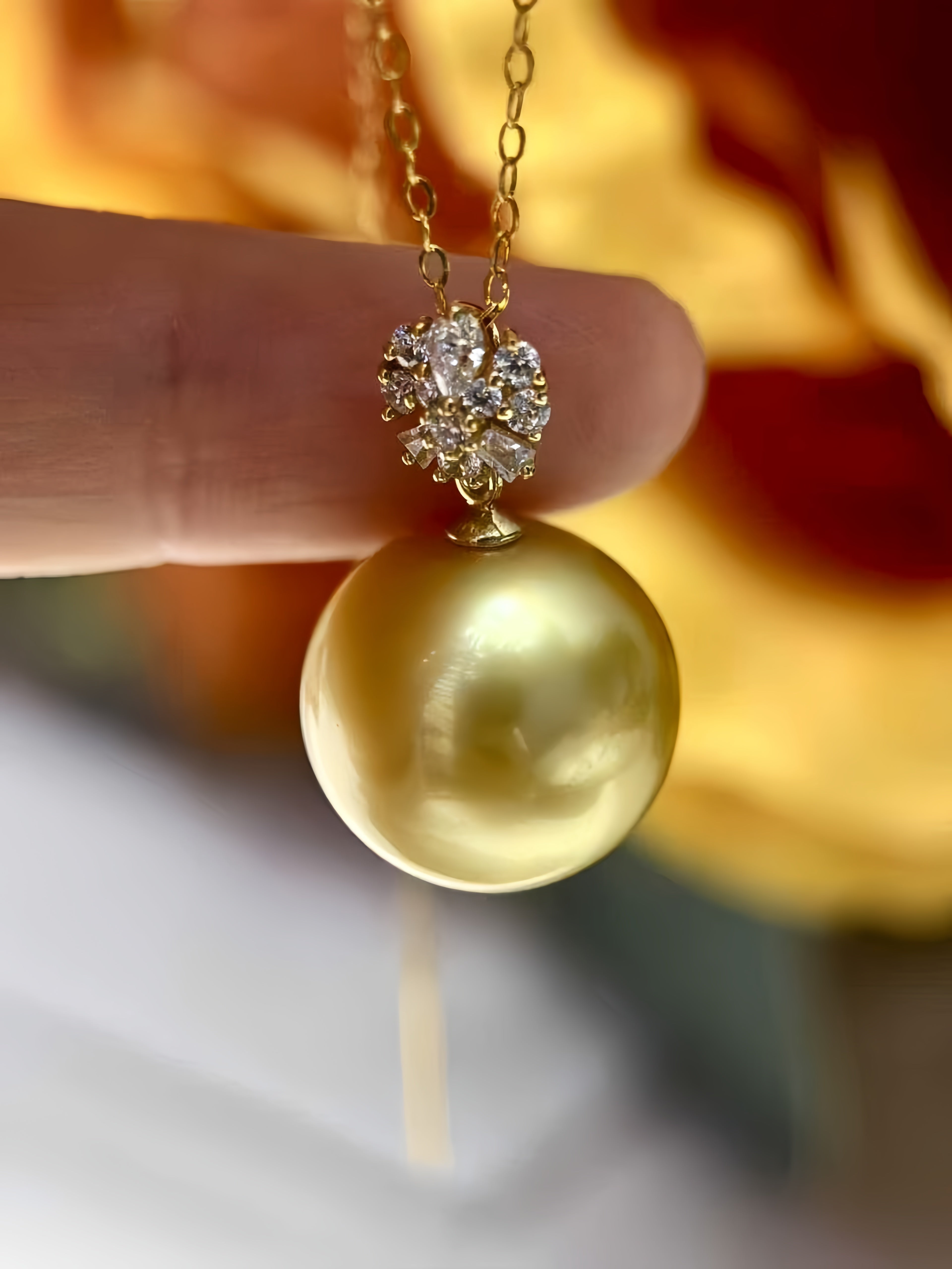 A close-up of a gold pendant necklace held by a finger, featuring a large round gold pearl pendant and a small cluster of clear stones above it, with a warm blurred background.