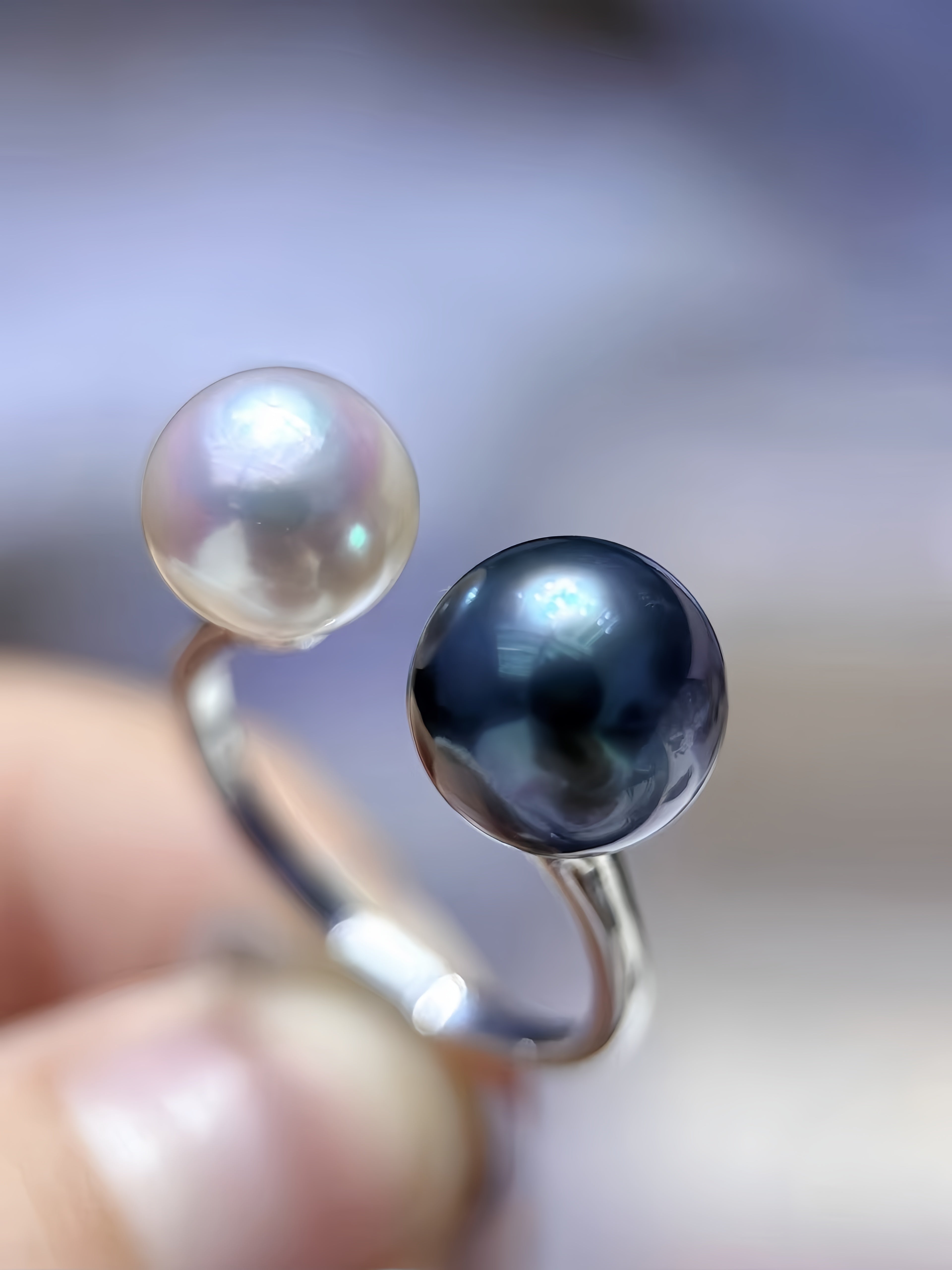 A close-up of a silver-toned ring held between fingers, featuring two round pearl-like beads: one lustrous off-white and one deep black or very dark navy.
