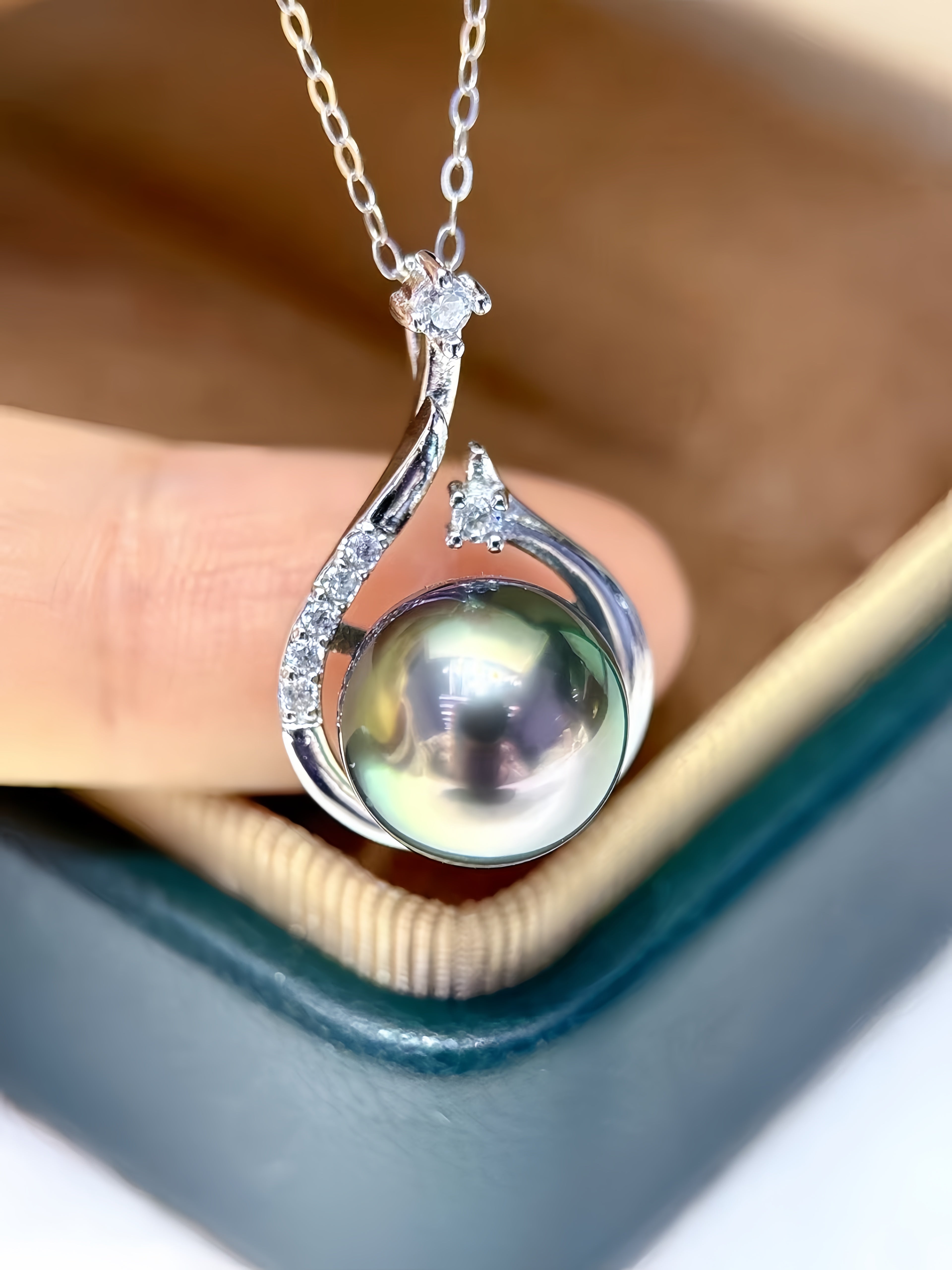 A silver pendant necklace featuring a large, round, iridescent greenish pearl accented with small clear stones, held by a hand over a jewelry box.